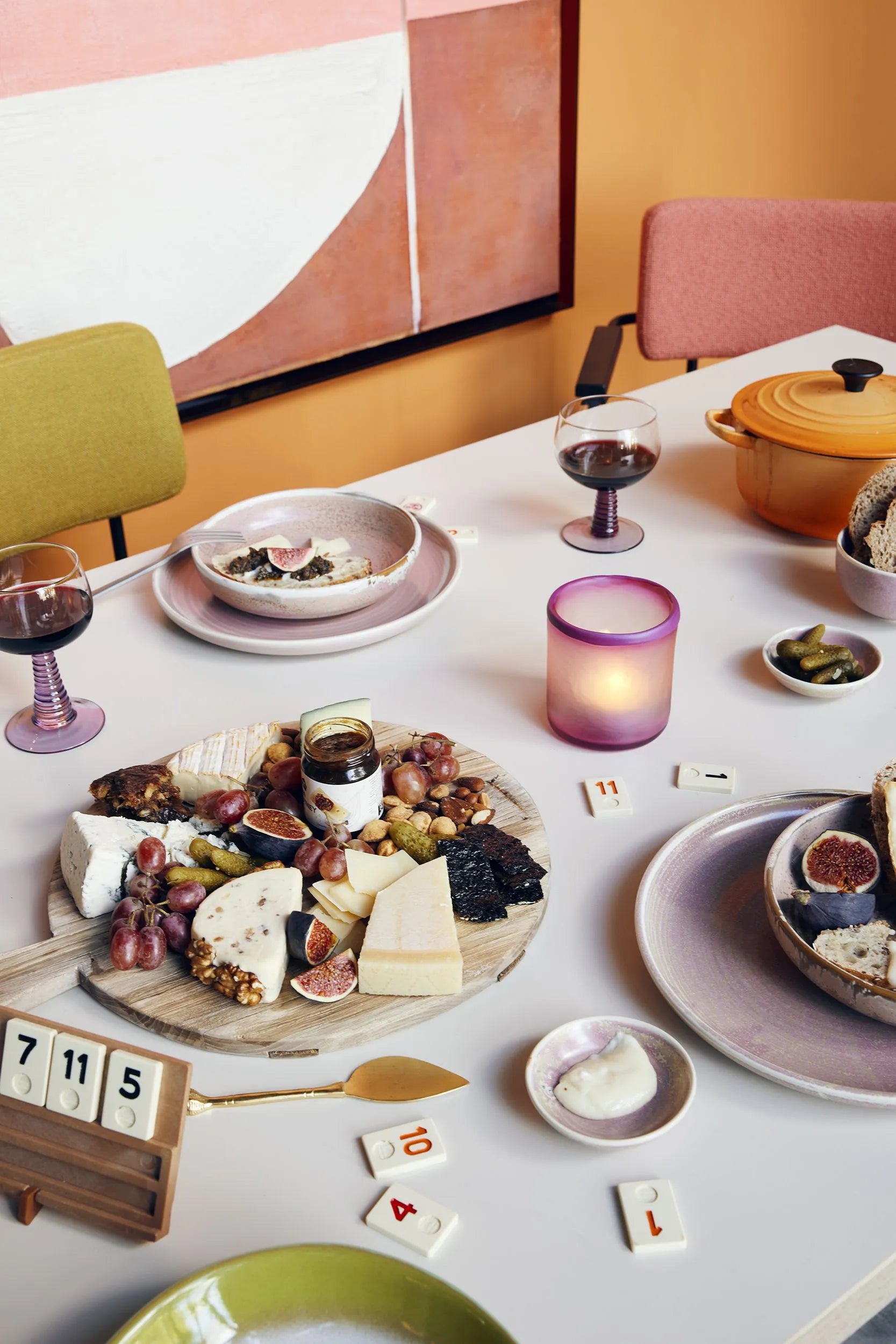 A table set for two features HKliving Chef Ceramics: Deep Plate M by HKliving, wine glasses, figs, a cheese and grape platter, candlelight, a yellow Dutch oven, a board game, and colorful chairs with abstract art on an orange wall.
