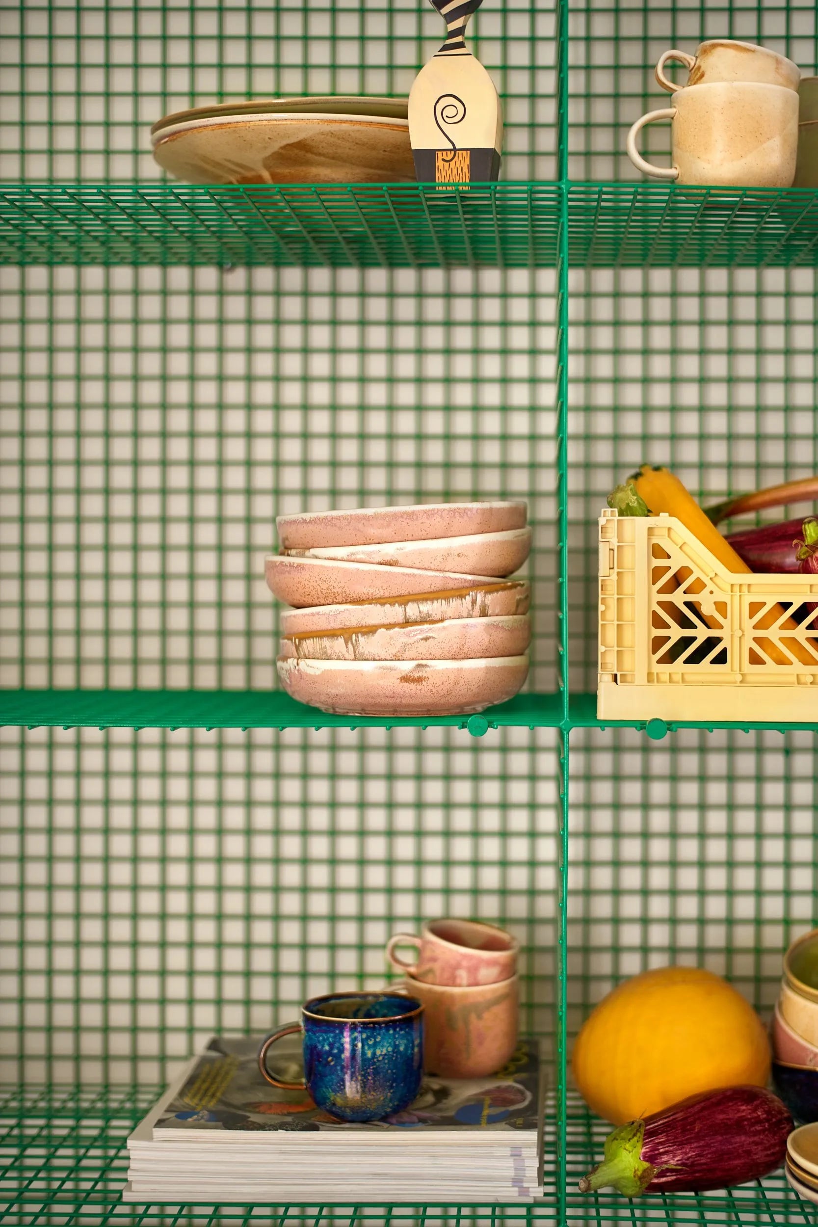 A green wire shelf displays HKliving Chef Ceramics: Deep Plate M by HKliving, stacked with bowls, mugs, and plates alongside a crate of vegetables, a yellow melon, and books, set before a charming green checkered wall.