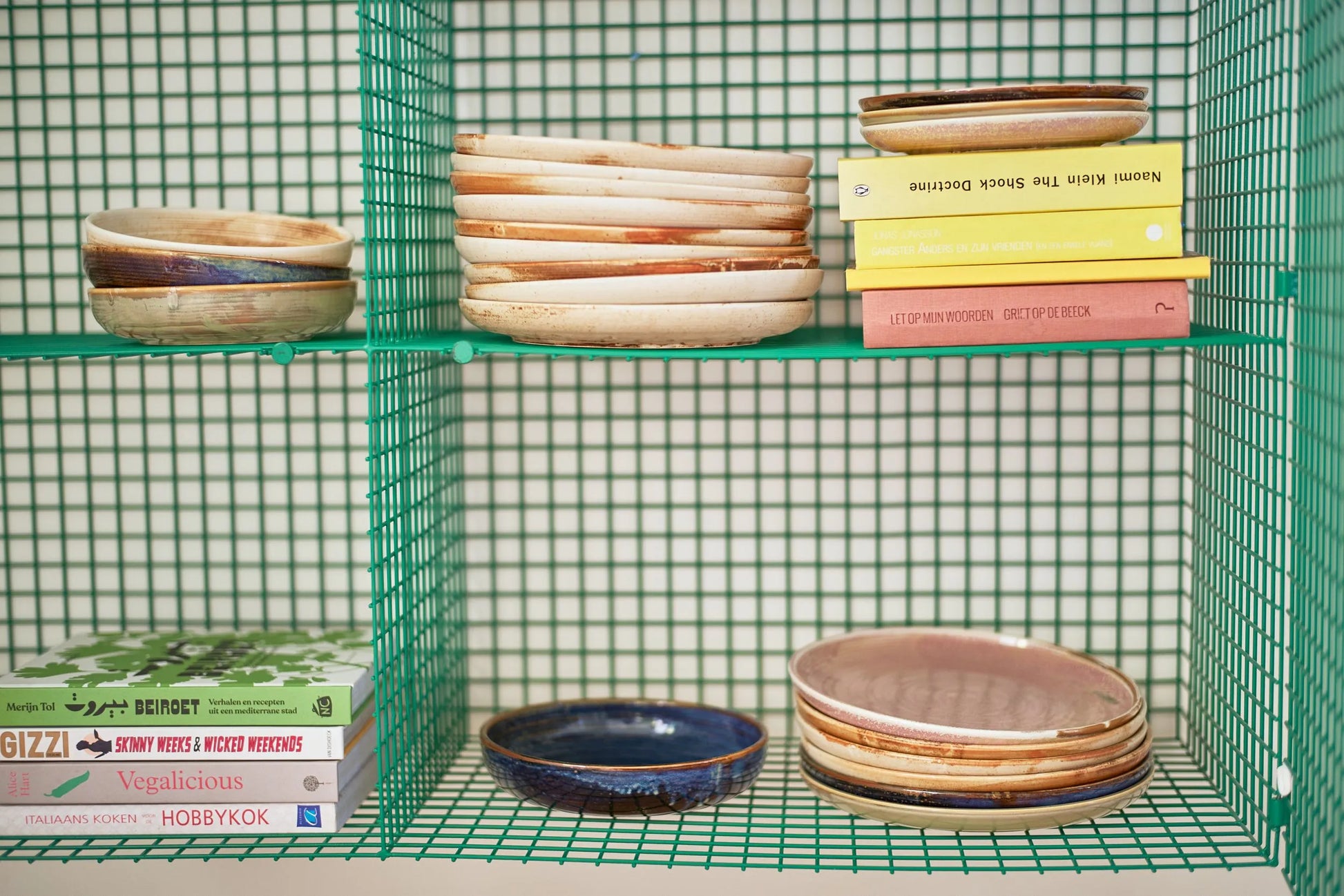 Green wire shelves neatly display stacks of HKliving Chef Ceramics: Dinner Plates and bowls, paired with vibrant cookbooks. The setup is tidy and modern, highlighting these stylish HKliving essentials for any kitchen.