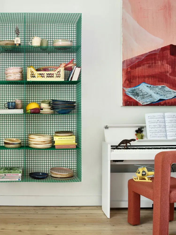 A green wire wall shelf with stacked bowls, cups, books, and an HKliving Chef Ceramics Dinner Plate sits by a white keyboard piano featuring sheet music, a dinosaur figure, toy car, orange chair, and an abstract painting above.