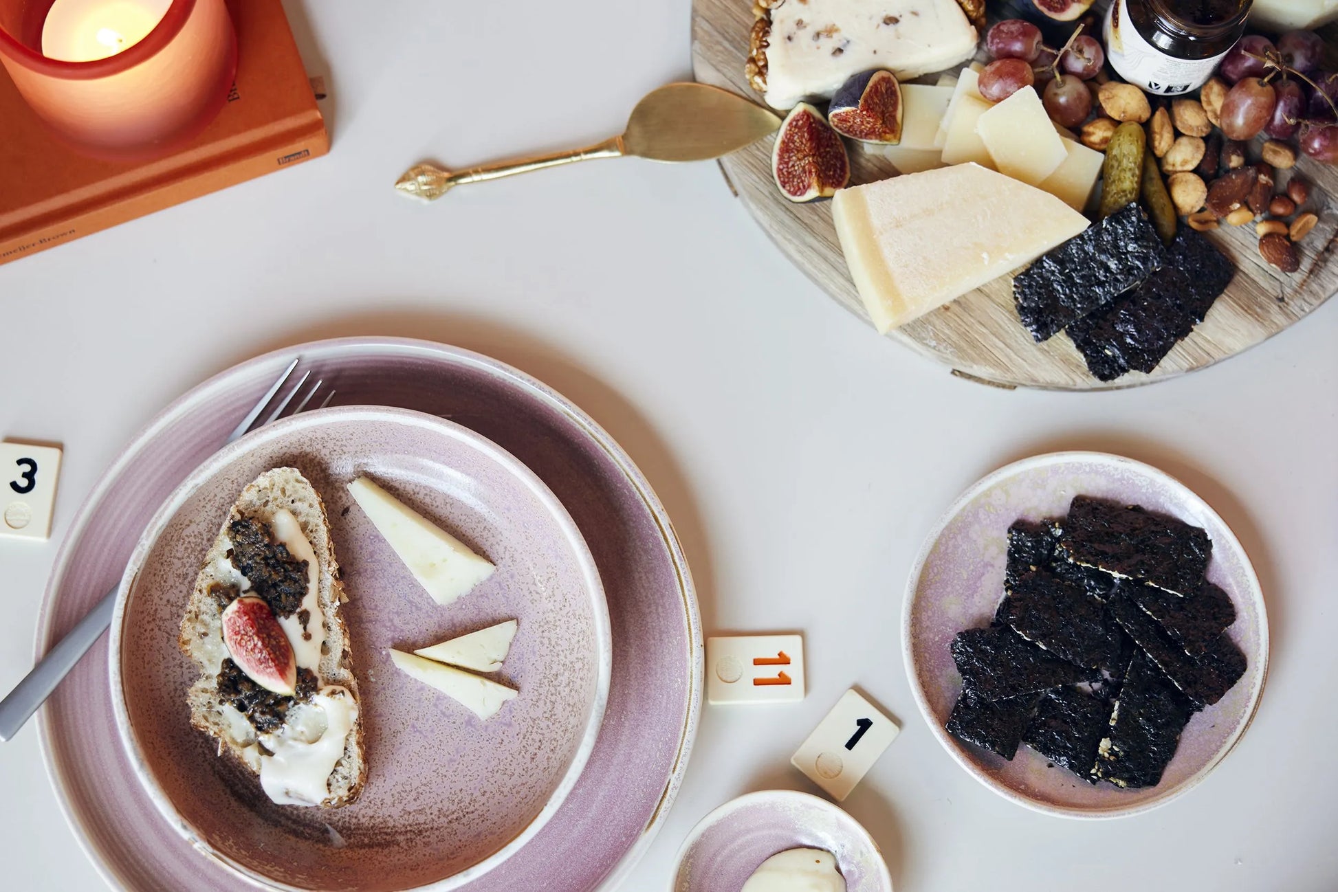 A table set with essentials features a HKliving Chef Ceramics: Dinner Plate holding cheese, crackers, figs, and seaweed snacks, complemented by a lit candle, gold cheese knife, and game tiles numbered 3 and 11.