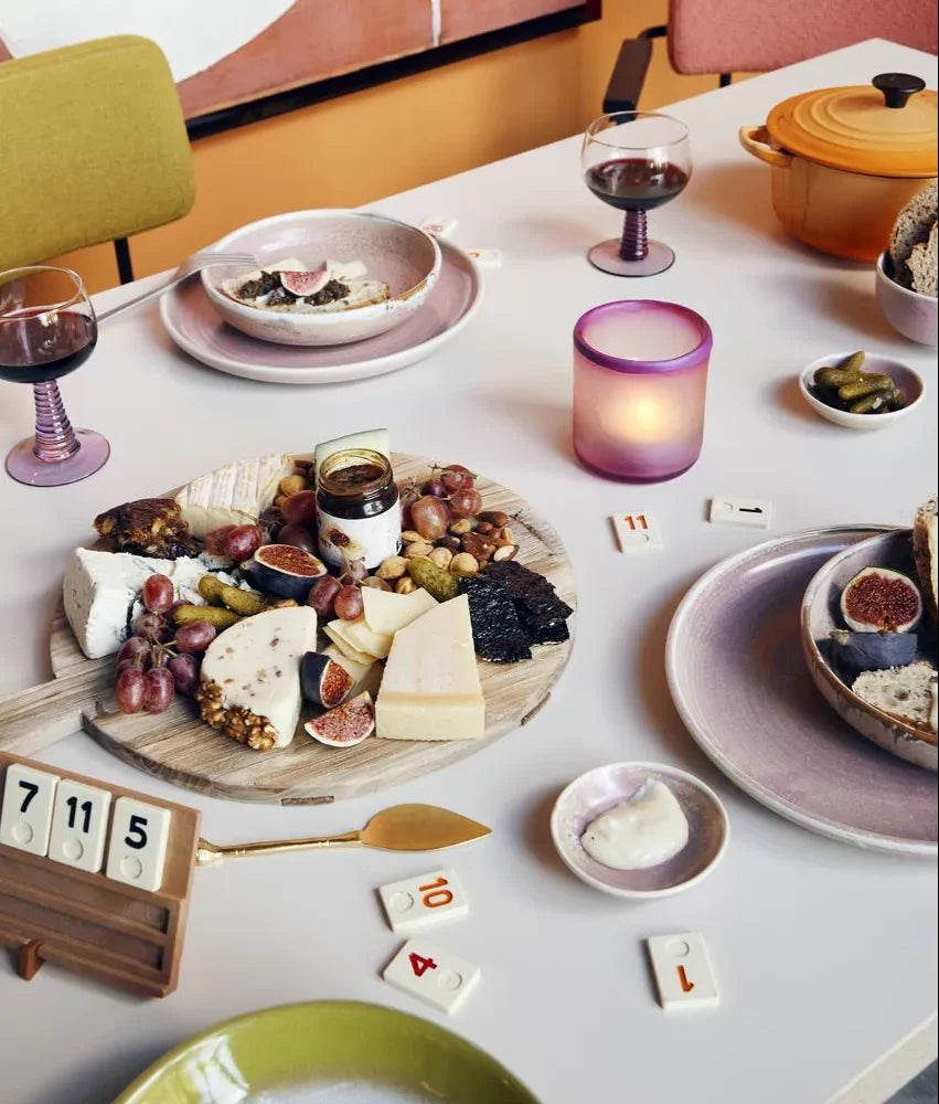 A meal table is set with wine, a cheese and fruit platter, figs on plates including the HKliving Chef Ceramics: Dinner Plate by HKliving, tableware, a lit candle, pickles, a bread pot, and scattered Rummikub tiles.