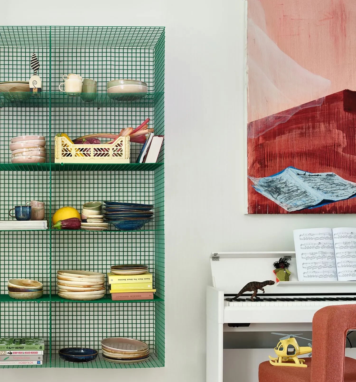 A green wire grid shelf displays HKliving Chef Ceramics Mugs and a yellow crate. To the right, there’s a white piano with sheet music, a toy dinosaur, and a modern red chair. An abstract red and blue painting hangs above.