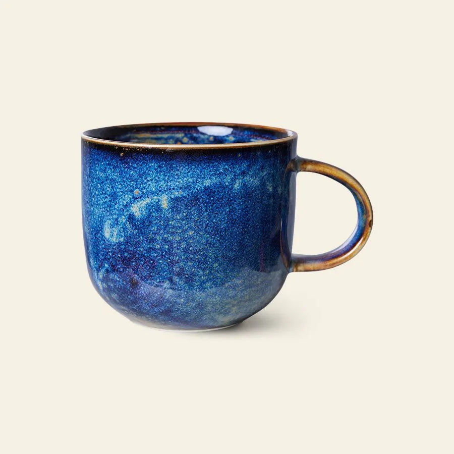 The HKliving Chef Ceramics: Mug by HKliving features a rounded, artisan-inspired design with a glossy deep blue speckled glaze and brown-tinted handle—an inviting tableware essential set against a light background.