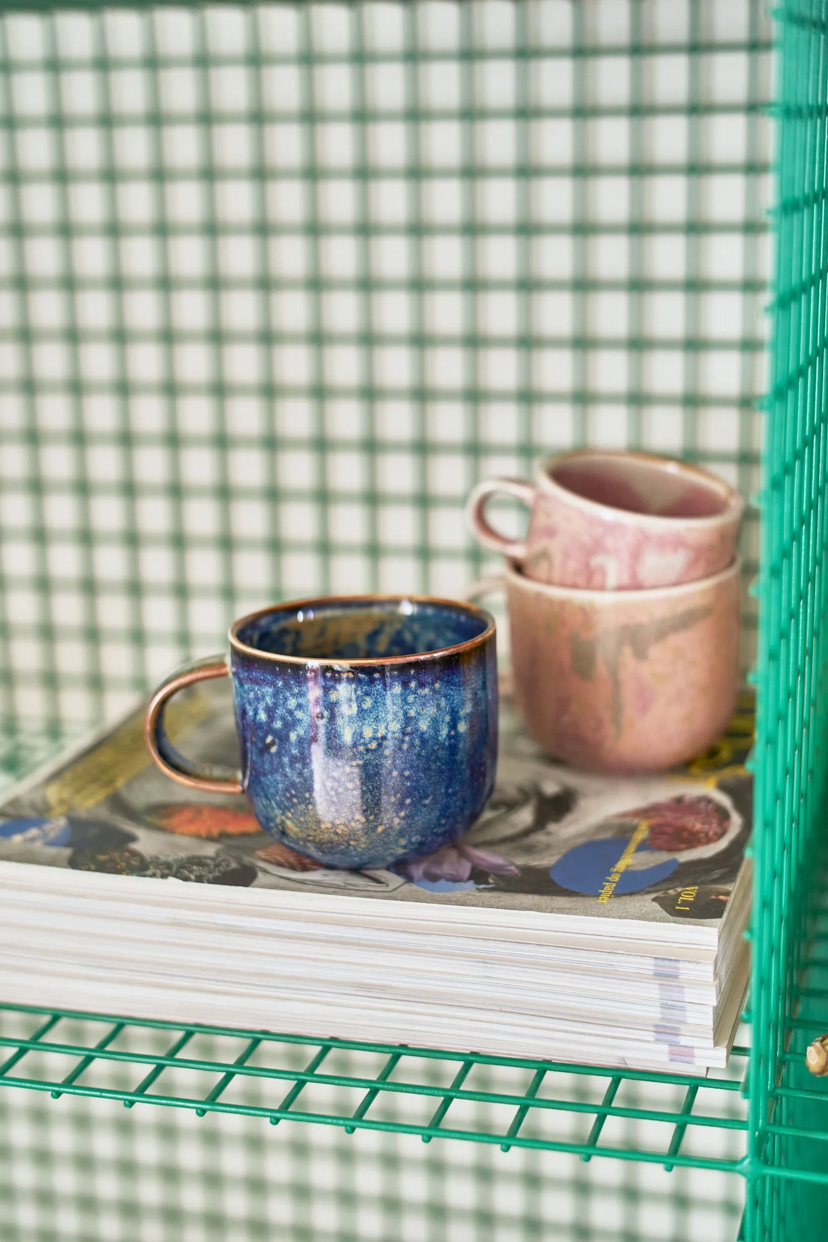 Three HKliving Chef Ceramics mugs—one blue, two pink—exude artisanal charm stacked on magazines atop a green wire shelf and a checkered backdrop. Stylish tableware essentials by HKliving.