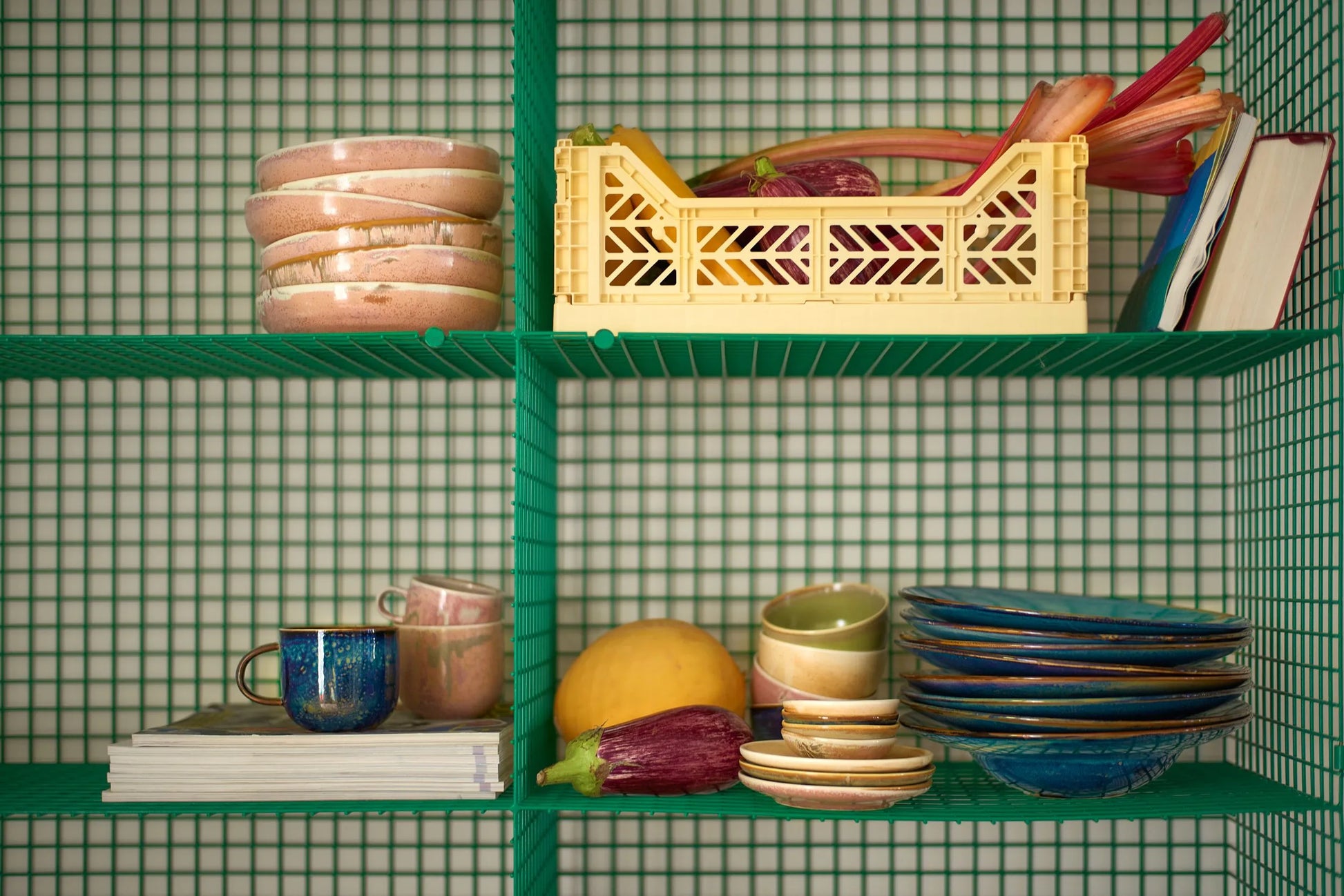 A green wire shelving unit displays HKliving Chef Ceramics: Mug along with stacked tableware, magazines, a yellow crate filled with vegetables, a book, yellow fruit, and two eggplants. A green grid background adds artisanal charm.