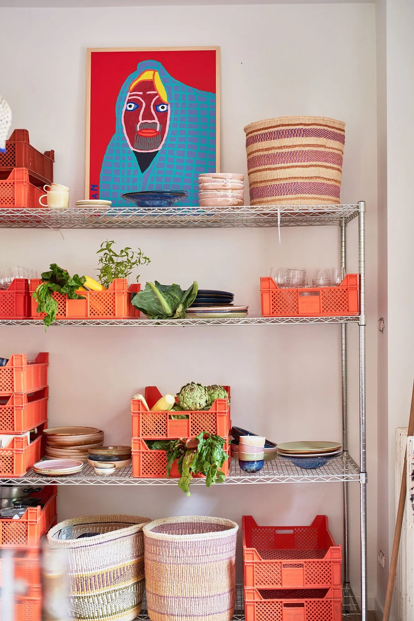 A metal shelving unit displays orange crates filled with HKliving Chef Ceramics: Pasta Plates, bowls, and fresh vegetables. Glassware and baskets rest on the shelves, with a vibrant portrait painting propped against the wall above.