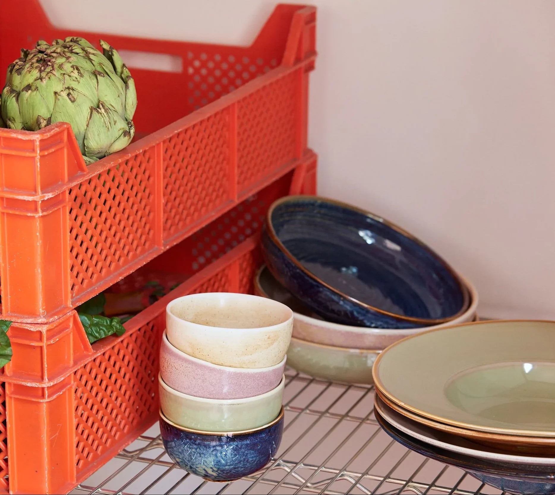 A red plastic crate with an artichoke sits on a metal rack beside a stack of HKliving Chef Ceramics tableware, including colorful bowls, plates, and the HKliving Chef Ceramics: Pasta Plate.