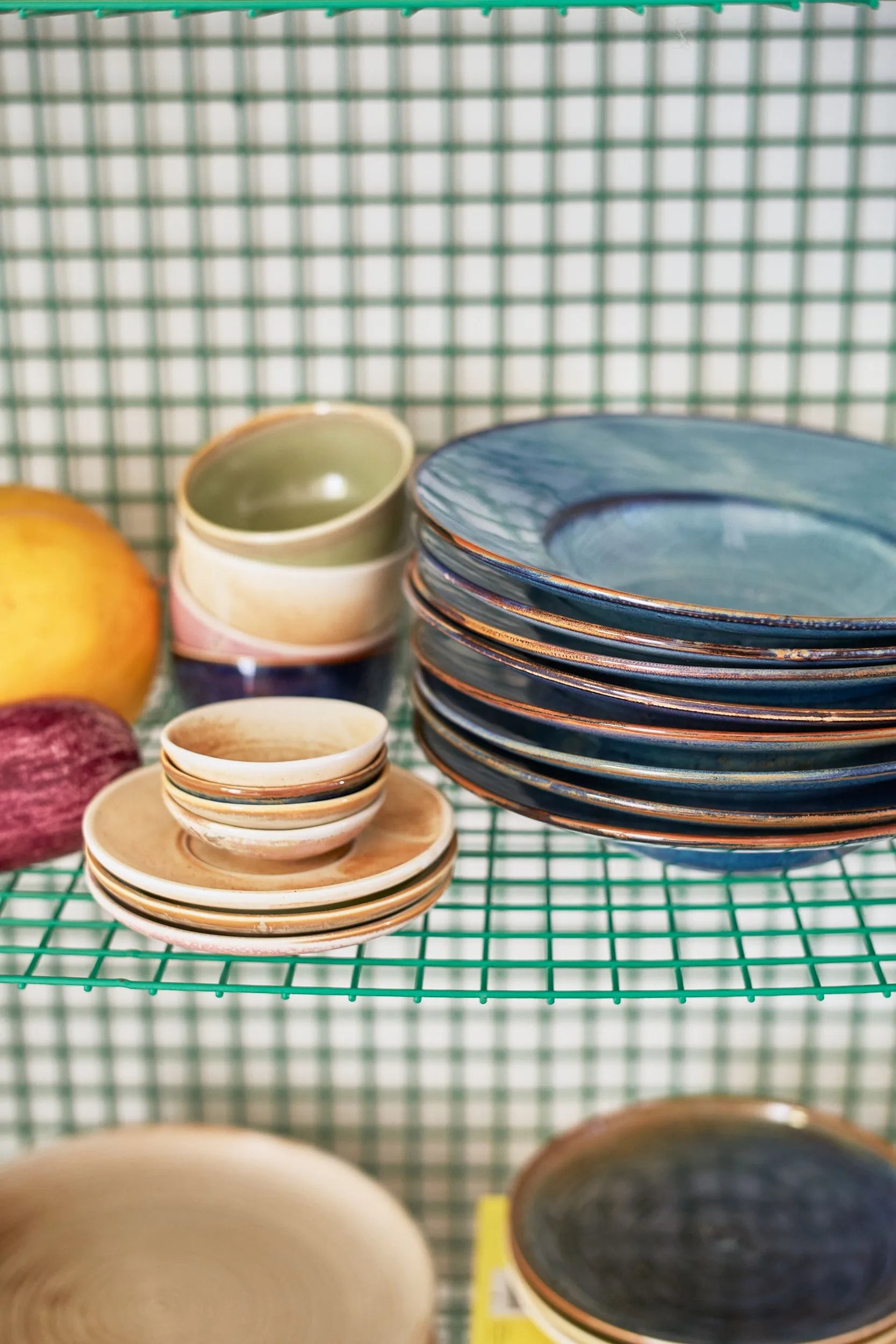 A stack of HKliving Chef Ceramics Pasta Plates sits on a green wire shelf beside a grapefruit and red onion, with a gridded tile wall behind—stylish tableware essentials from HKliving for any kitchen.