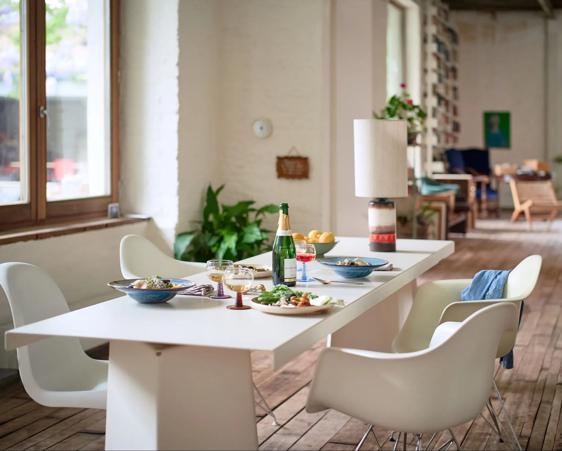 A modern dining room features a white rectangular table set with HKliving Chef Ceramics: Pasta Plates by HKliving, wine glasses, and essentials. Four white chairs surround the table atop wooden floors with large windows in the background.