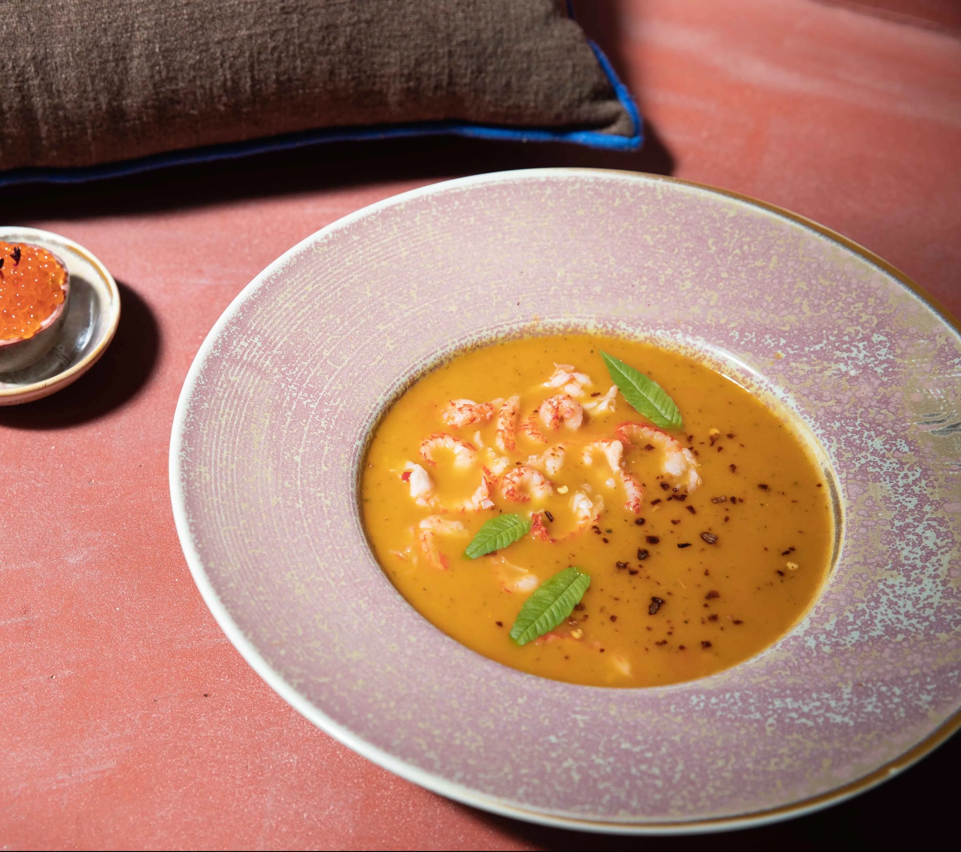A bowl of creamy orange soup with seafood and herbs is served in an HKliving Chef Ceramics: Pasta Plate by HKliving, set on a pink table with a small tart, brown napkin, and tableware essentials.
