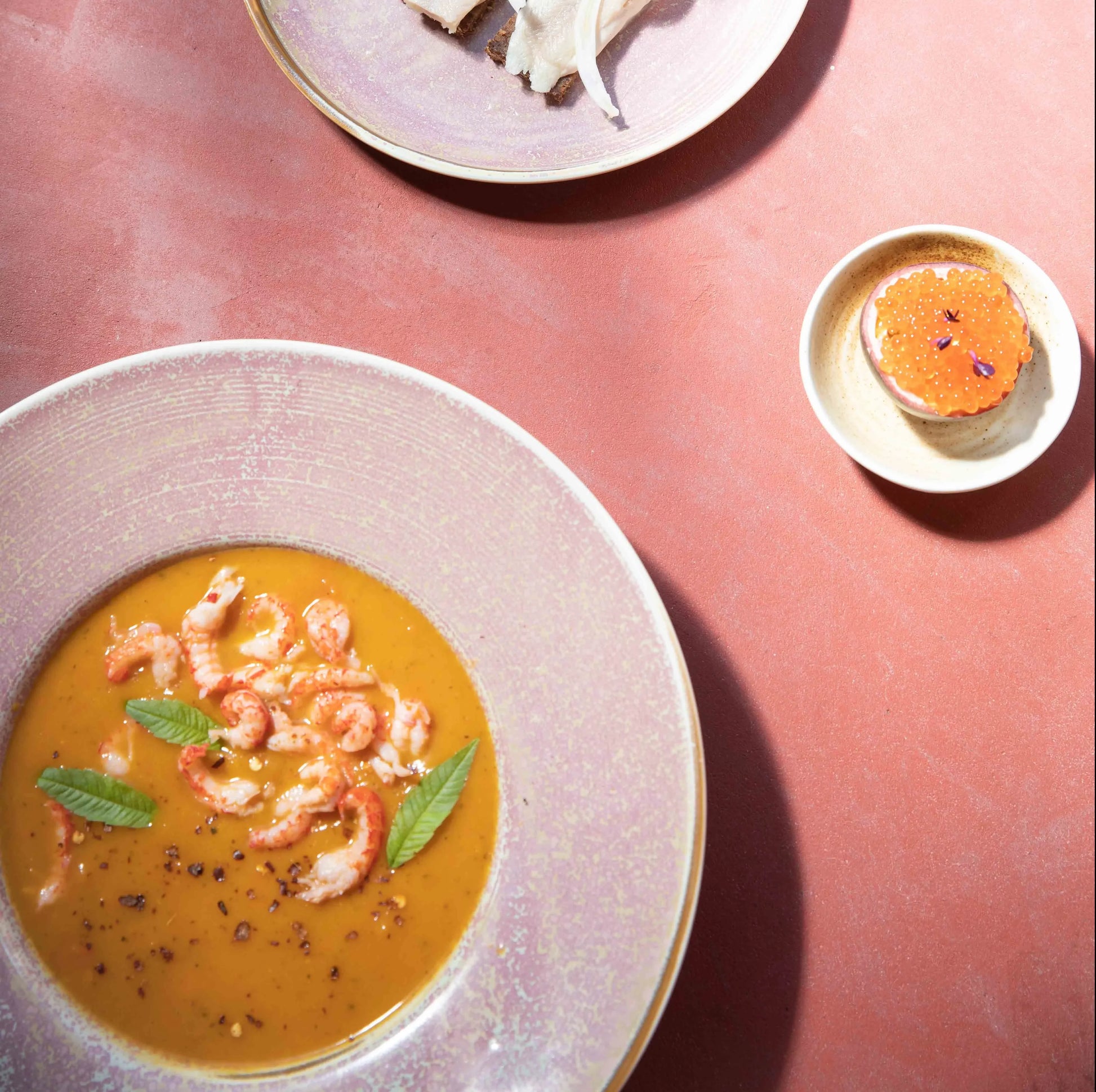 A bowl of creamy orange soup with shrimp and herbs is served in HKliving Chef Ceramics: Pasta Plate, accompanied by sliced bread and an orange-topped pastry, all displayed on a reddish surface.