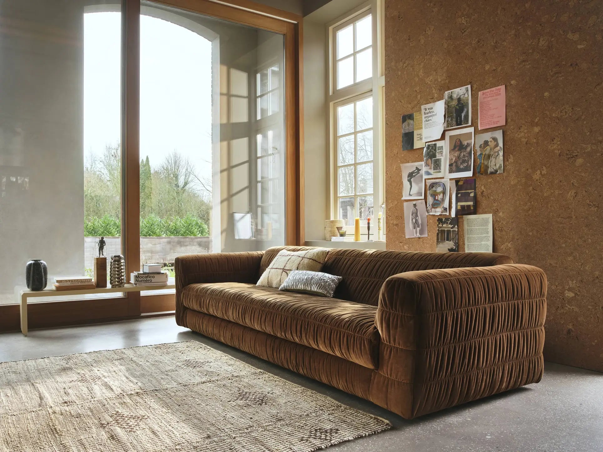 A cozy living room with the HKliving Club Couch in brown velvet, featuring pleated upholstery, a textured beige rug, a cork wall with photos and art, and tall windows bringing in natural light and views of greenery.