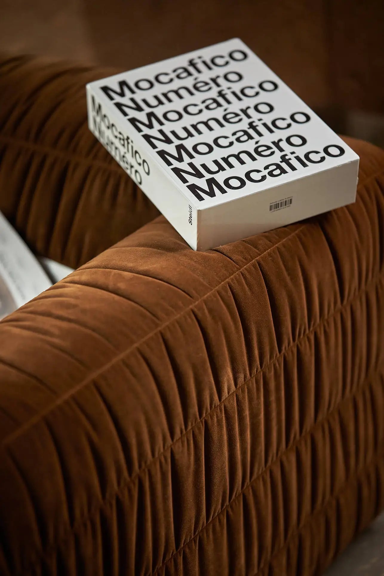 A white book with bold black text reading Mocafico Numéro lies on the HKliving Club Couch by HKliving, its velvety pleated upholstery enhancing the scenes sense of comfort.