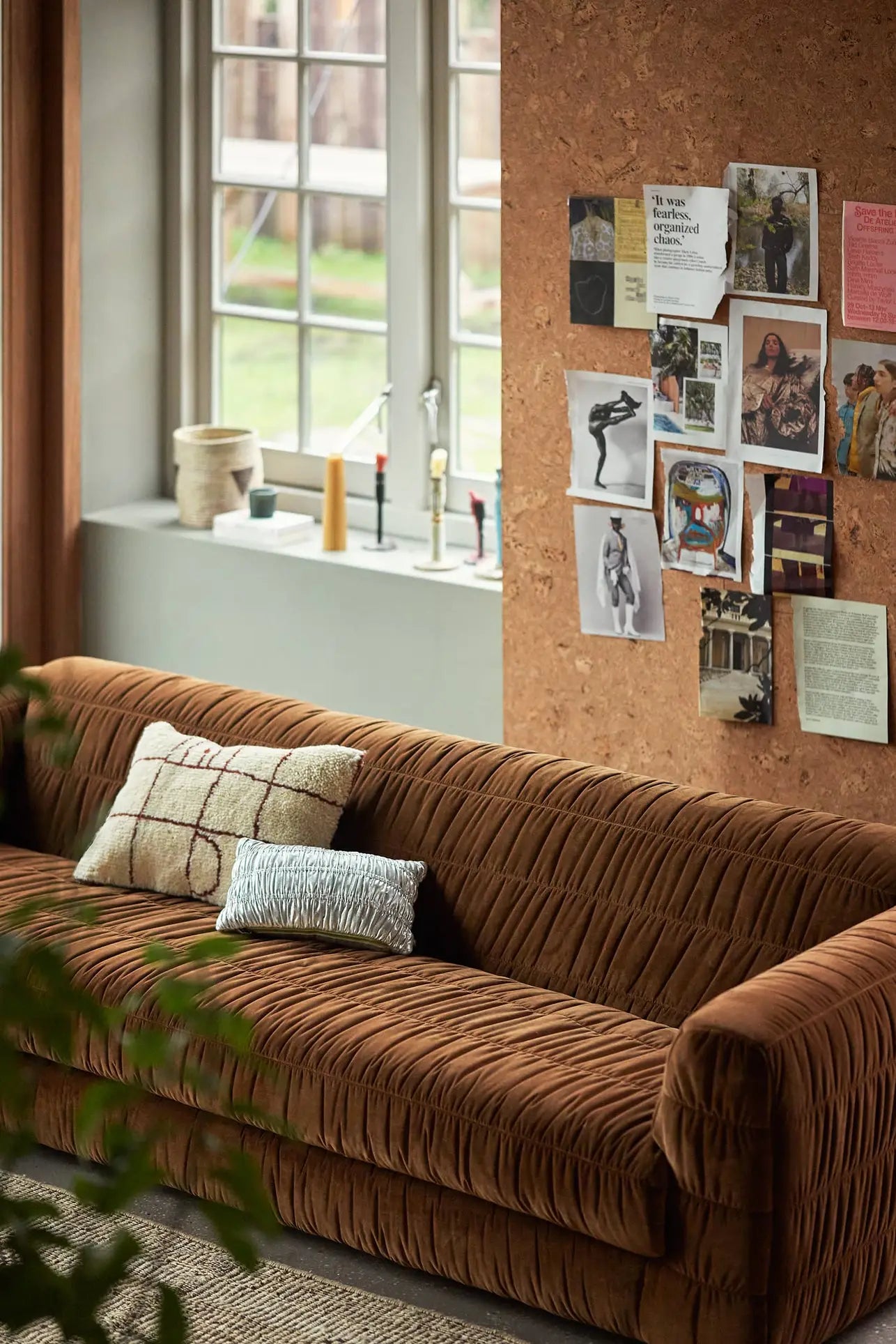 A cozy living room with an HKliving Club Couch in brown velvet, featuring plush seating, two pillows, and a corkboard wall with photos. The windowsill displays pottery and candles as sunlight pours through large windows.