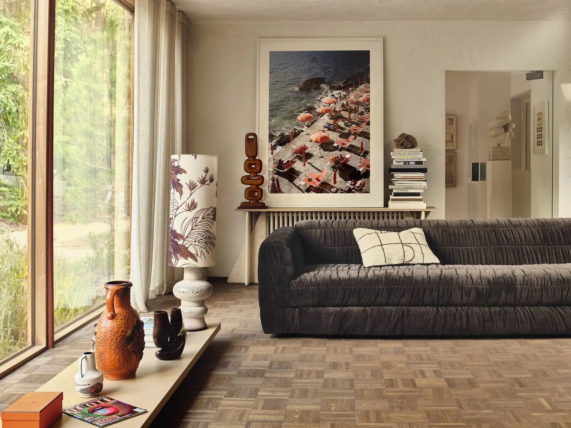 A cozy living room with the HKliving Club Couch in dark velvet and pleated upholstery, a modern lamp, stacked books, vases on a low table, sheer curtained window, and an aerial beach photo above the console.
