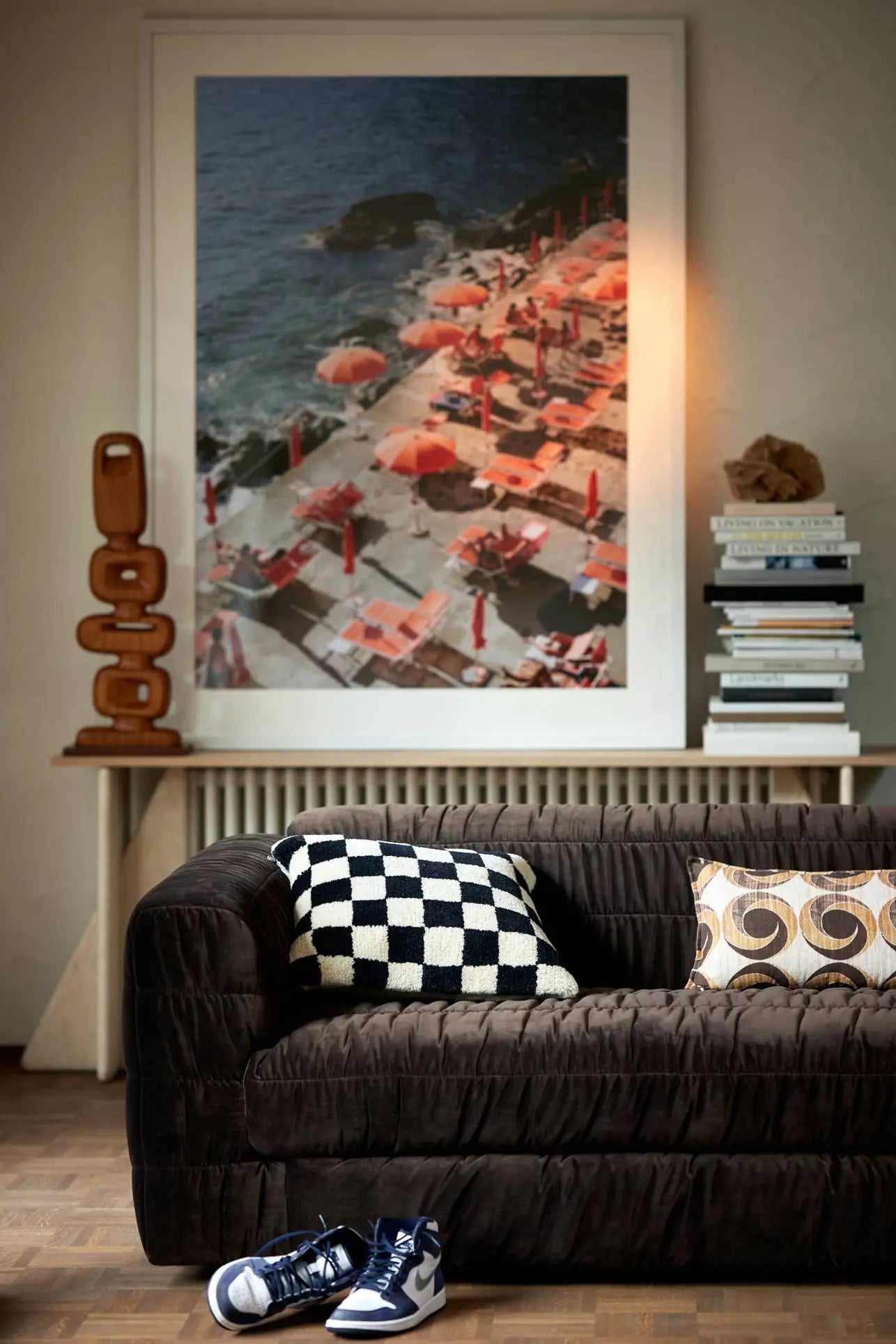 A cozy living room with an HKliving Club Couch in dark velvet and pleated upholstery, two patterned pillows, sneakers on the floor, and a large seaside photo with orange umbrellas and lounge chairs above the radiator.