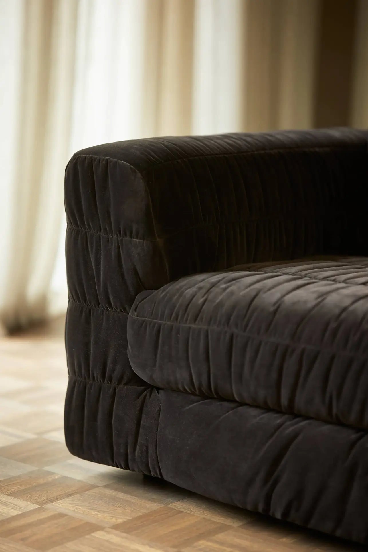 The HKliving Club Couch features plush, dark upholstery with elegant pleats on the armrest and seat, illuminated by soft natural light through sheer curtains and set atop a warm wooden parquet floor for inviting comfort.