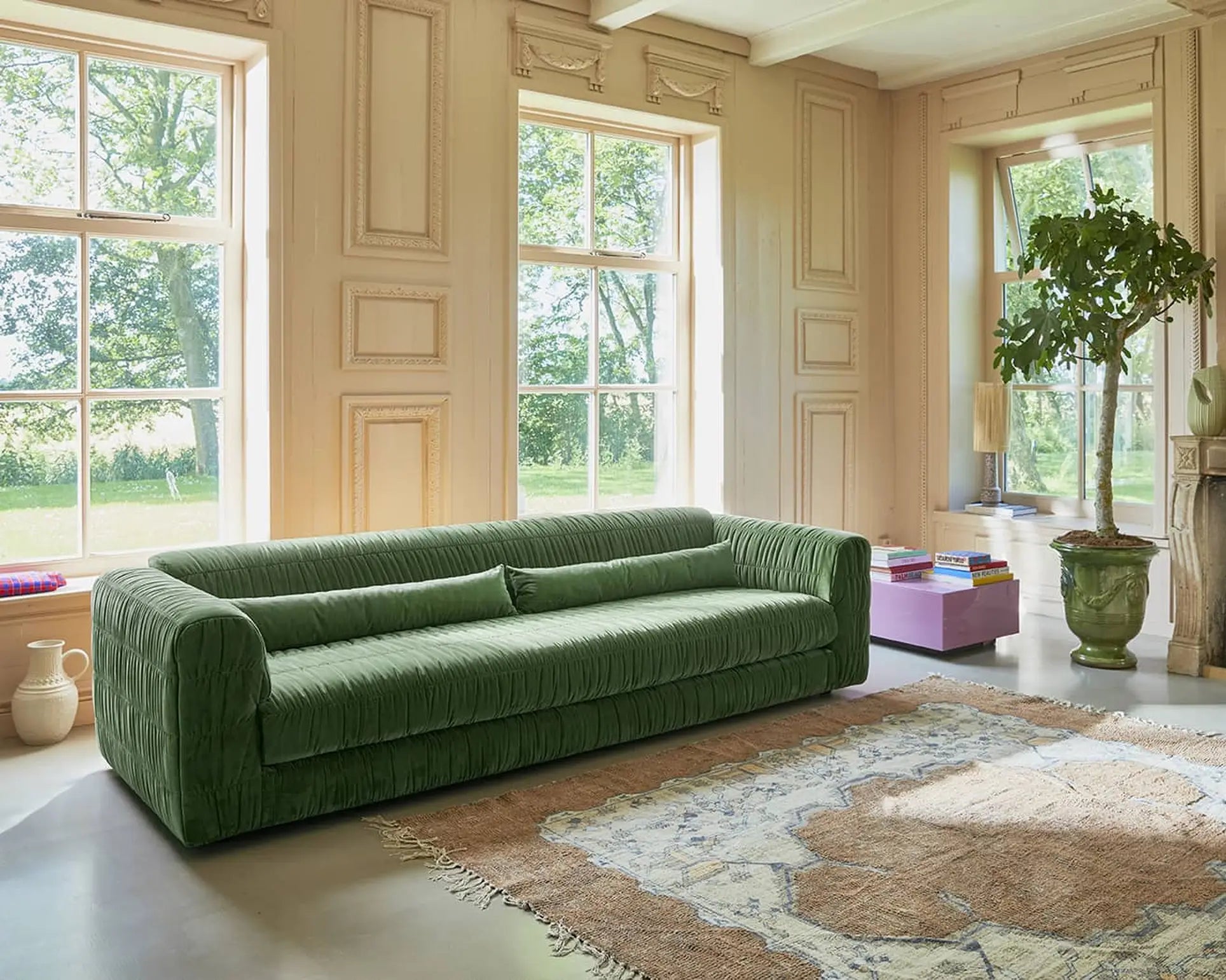 A spacious living room with large windows, beige paneled walls, an HKliving Club Couch in green pleated upholstery, a patterned rug, a purple coffee table, houseplants, and decorative objects filled with natural light.
