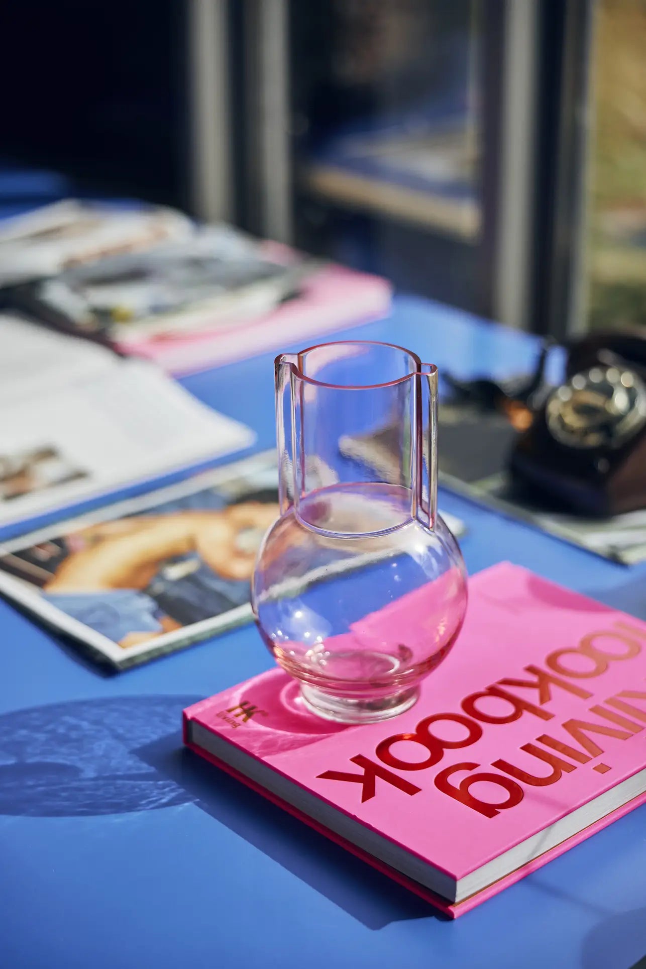 The HKliving Glass Vase Sundae Pink sits atop a bright pink book on a blue table, with magazines and a black rotary phone in the background near the window.