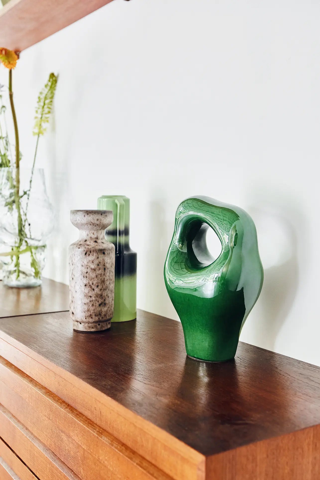 Three HKliving HK Objects: Ceramic Sculpture vases—a speckled beige, a green glass, and a glossy green sculptural vase—add modern decorative accents to a wooden surface, with blurred glass vases and flowers in the background.