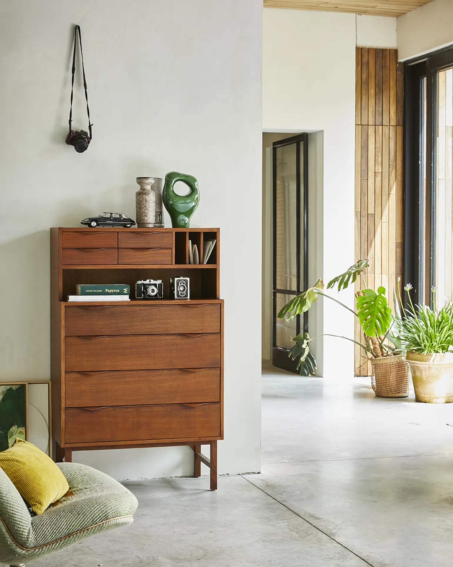 A modern, light-filled room features a mid-century wooden dresser topped with vintage cameras, decor, and the HKliving HK Objects: Ceramic Sculpture by HKliving. Nearby sit plants in woven baskets, a green pillow on the floor, and a hanging camera as décor.