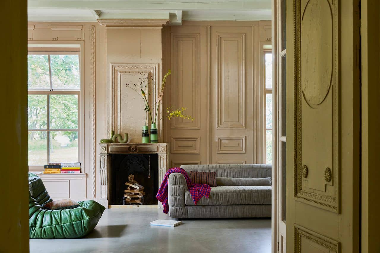 A cozy living room with beige paneled walls, a gray sofa with a pink blanket, a green lounge chair, and a fireplace adorned with plants and the HKliving HK Objects: Ceramic Twisted Vase, adding sculptural style by HKliving.