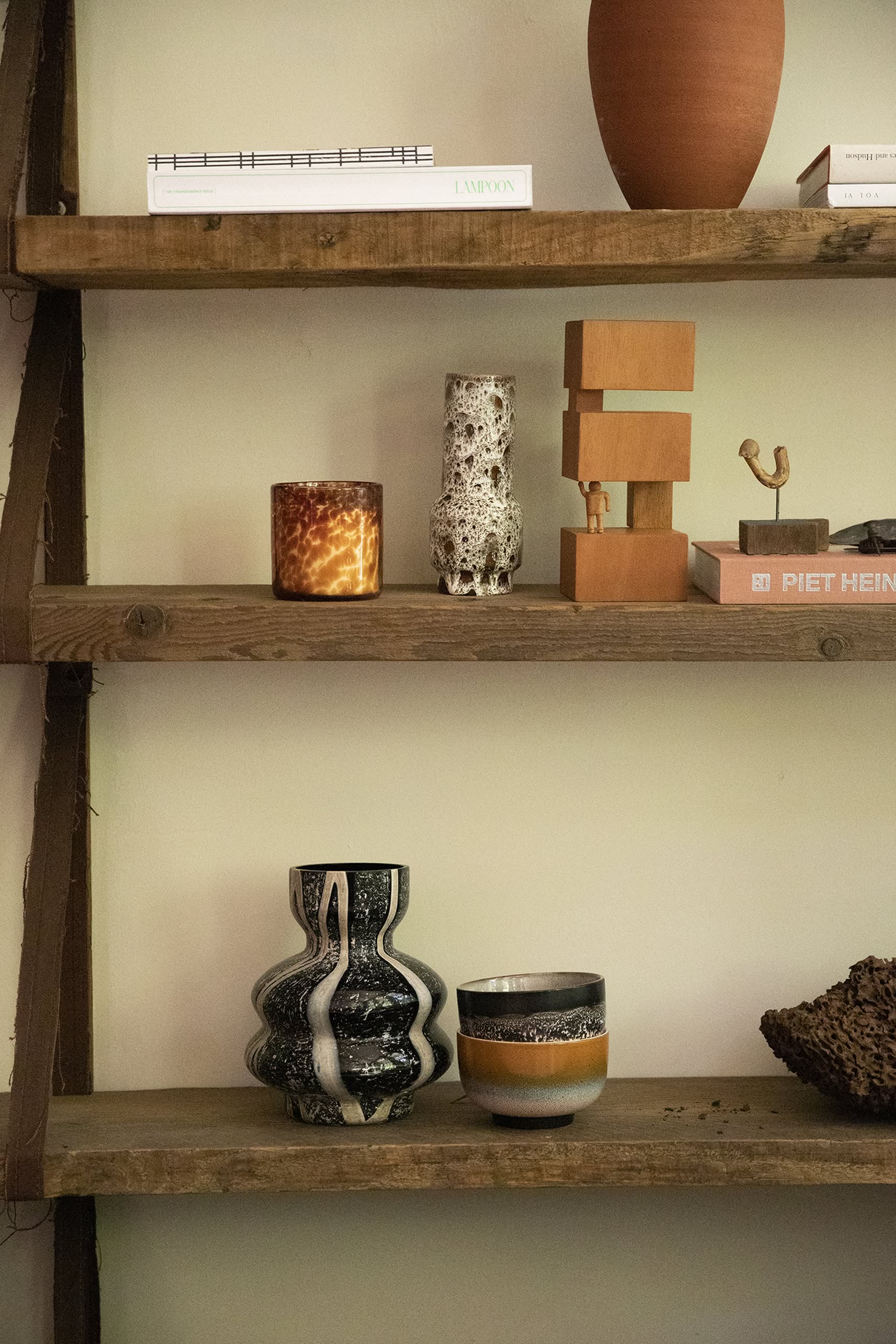 HKliving HK Objects: Empowered by HKliving—featuring a candle, textured and patterned vases, wooden blocks, books, and a hand-carved figure—are displayed on shelves against a light-hued Bauhaus-inspired wall.