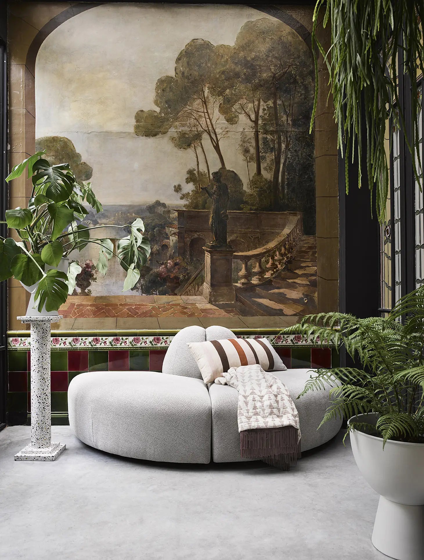 The HKliving Jax Couch, Angle in modern gray sits with a striped fringed throw before a classical mural of trees and terrace. The room includes large plants, decorative tile accents, and striking arrangements for added elegance.