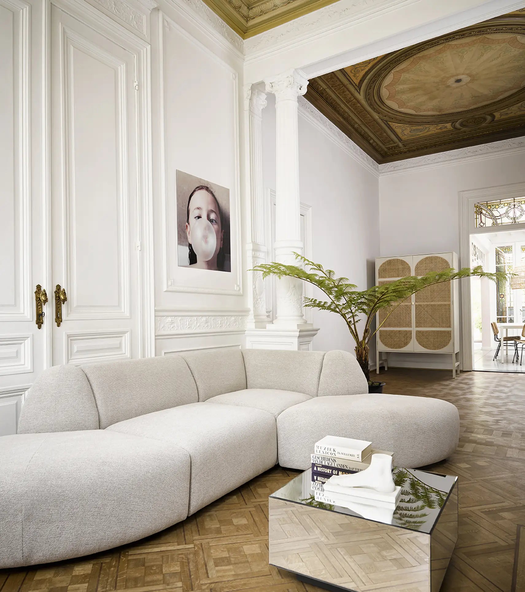 A stylish living room features the HKliving Jax Couch, Angle by HKliving, paired with a mirrored coffee table, curated books and plants. Ornate white walls, tall columns, and bubblegum art photography complete the versatile design.