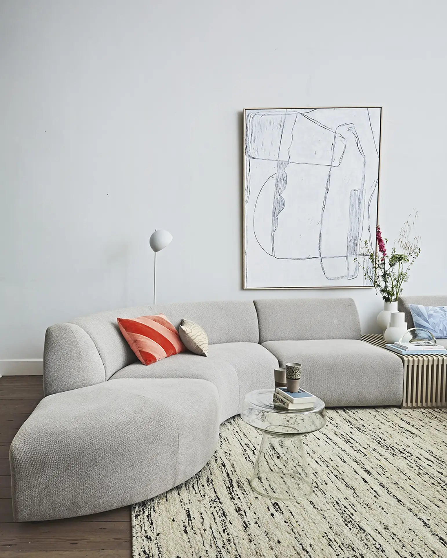 Modern living room featuring the HKliving Jax Couch, Angle by HKliving in light gray, a white lamp, round glass coffee table, framed abstract art, flowers on a striped side table—showcasing versatile design.