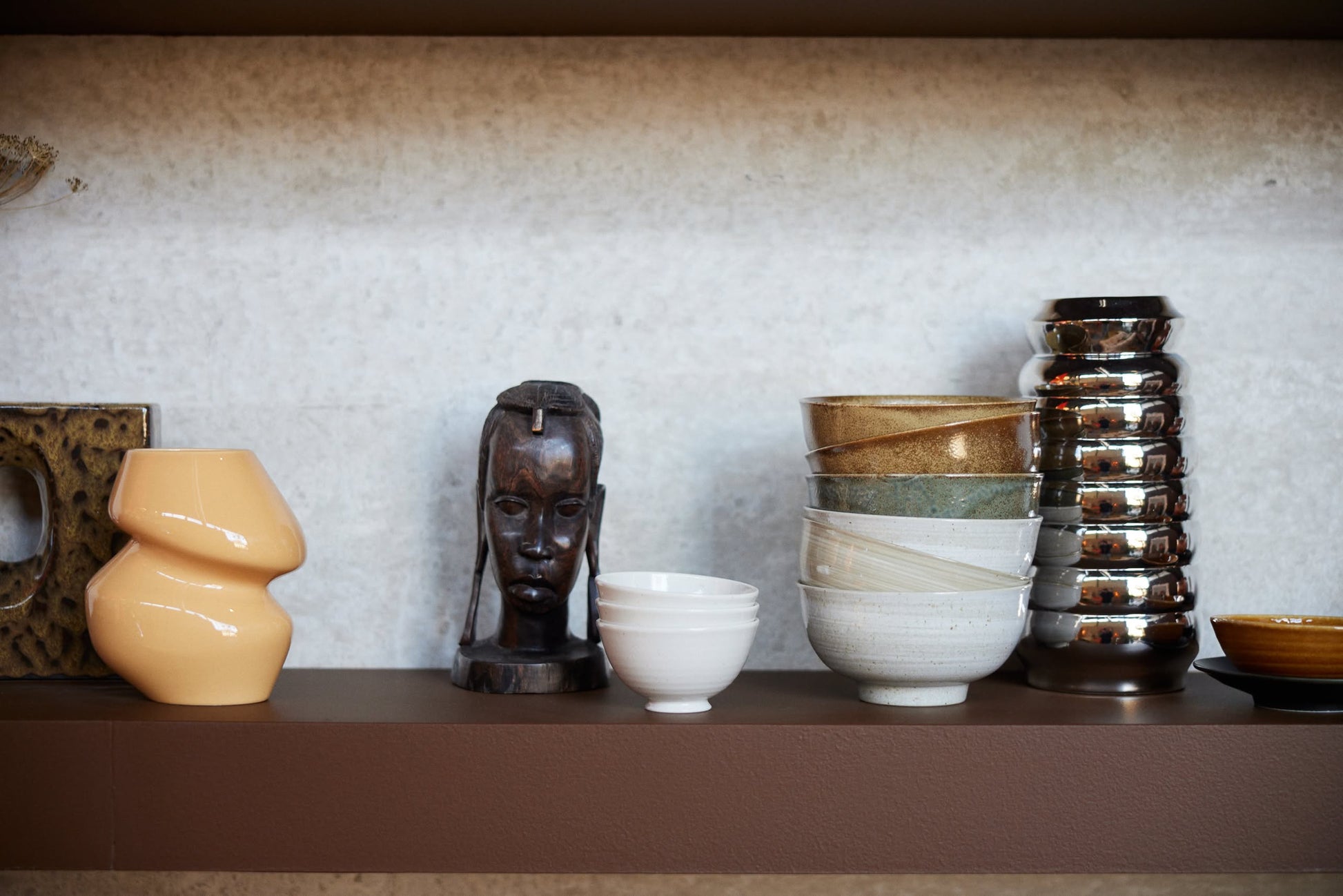 A shelf displaying a beige abstract vase, a dark carved wooden head sculpture, HKliving Kyoto Ceramics: Japanese Noodle Bowls, Set of 4 by HKliving in neutral tones, and several shiny metallic cups.