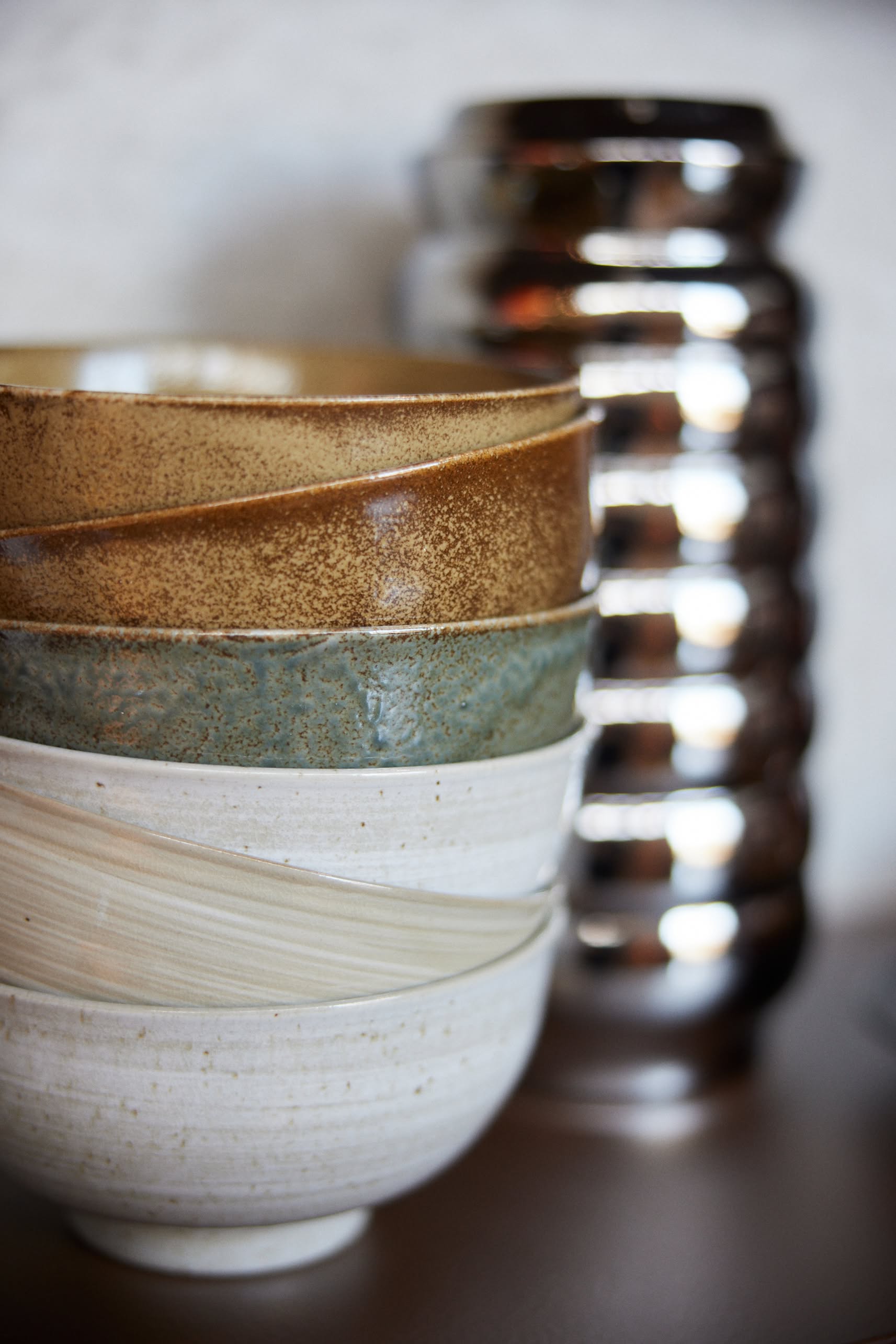 A close-up of three stacked HKliving Kyoto Ceramics Japanese Noodle Bowls (set of 4) in earthy tones sits before a tall, textured silver vase, with a softly blurred background.