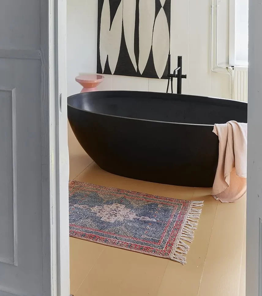 A modern bathroom features a matte black freestanding bathtub, a pink towel draped over the edge, the HKliving Printed Bathmat Overtufted on the pale floor, a pink bowl on a stand, and striking abstract black-and-white wall art.