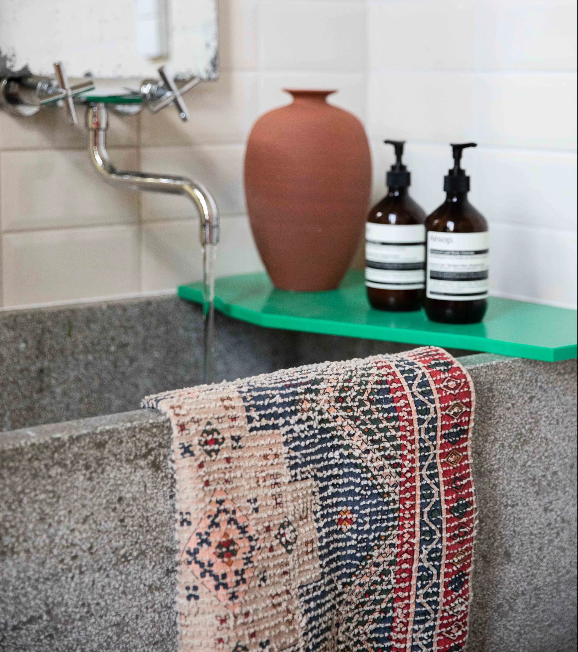 A close-up of a concrete sink with water running from a wall-mounted faucet features an HKliving Printed Bathmat Overtufted draped over the front and stylish decor—a green shelf with a brown vase and two pump bottles—in the background.