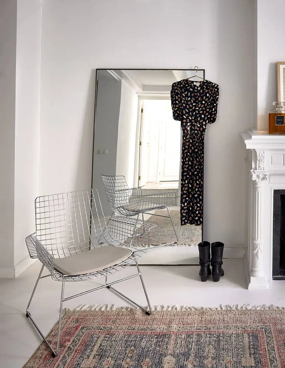 A black floral dress on a hanger and black boots sit before a large mirror near a white fireplace; in the foreground, an HKliving Printed Rug Overtufted (150x240) by HKliving and a modern wire chair with cushion add stylish touches.