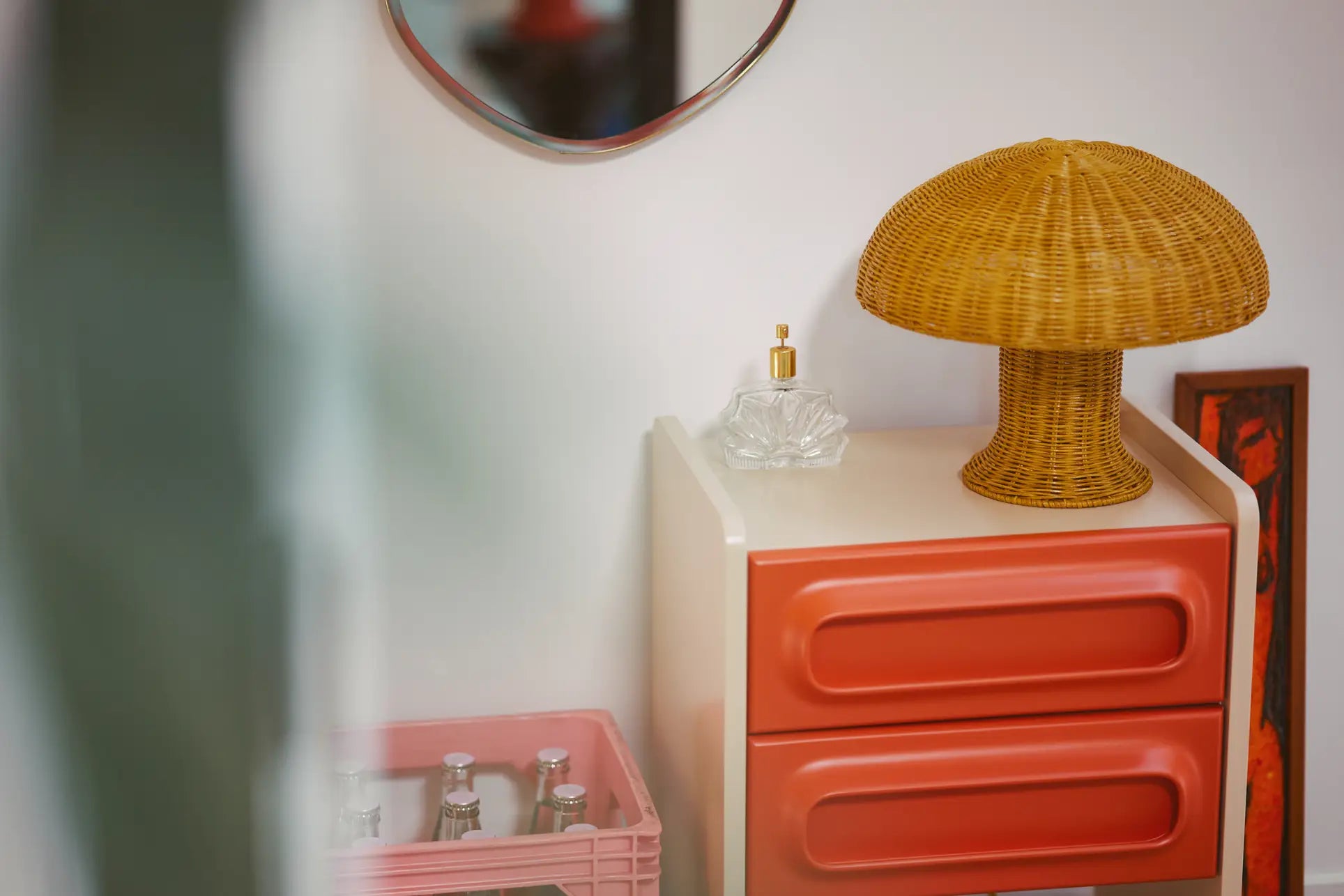 A retro-style room corner with country vibes, featuring a small white and orange cabinet, the HKliving Rattan Table Lamp by HKliving in yellow mushroom shape, cozy mood lights, a glass perfume bottle, a pink crate with bottles, part of a mirror, and wall art.