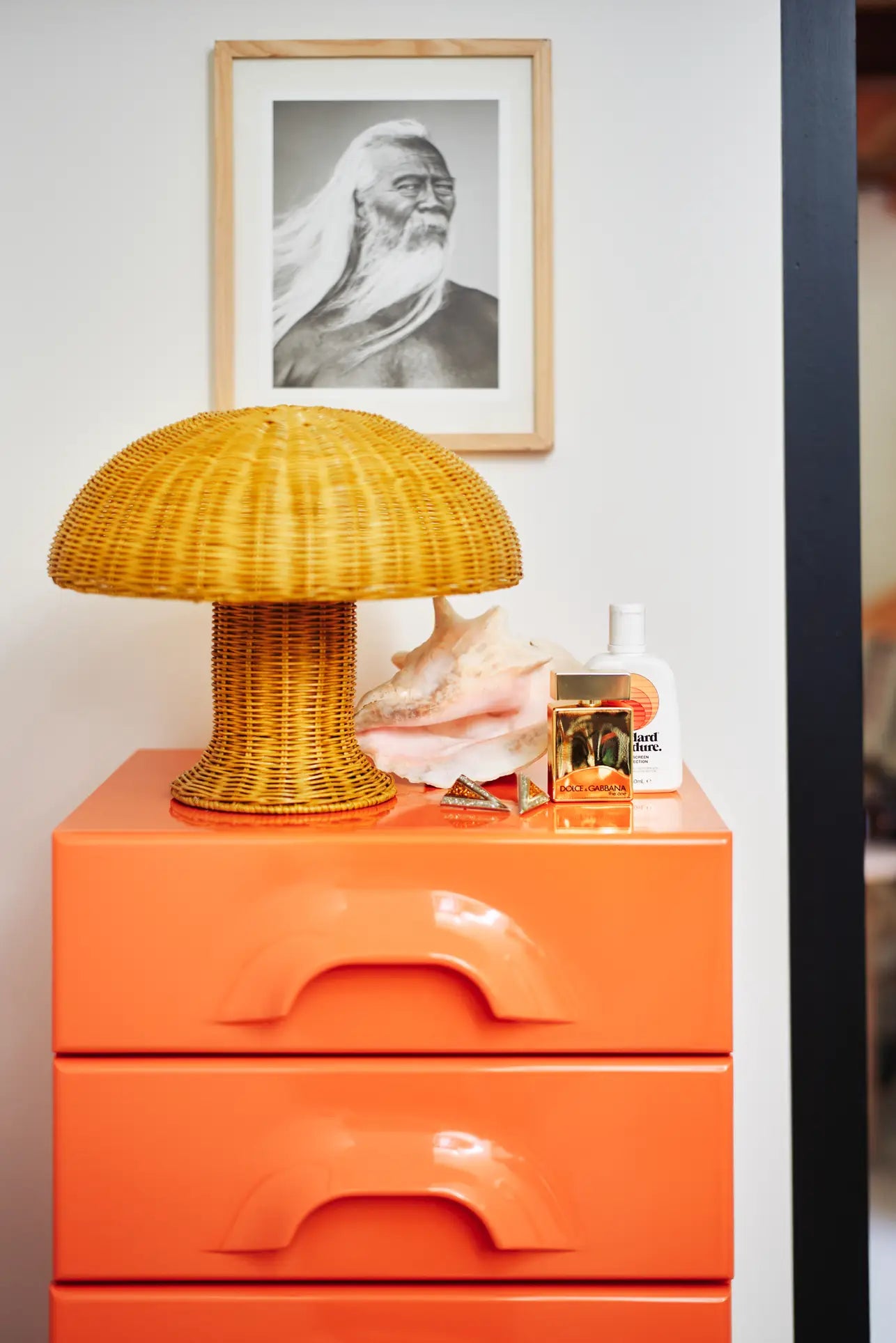 A bright orange dresser with curved handles displays the HKliving Rattan Table Lamp, casting a cozy glow, beside a large seashell, perfumes, and lotion. Above, a framed black-and-white photo of a bearded man with long hair decorates the wall.