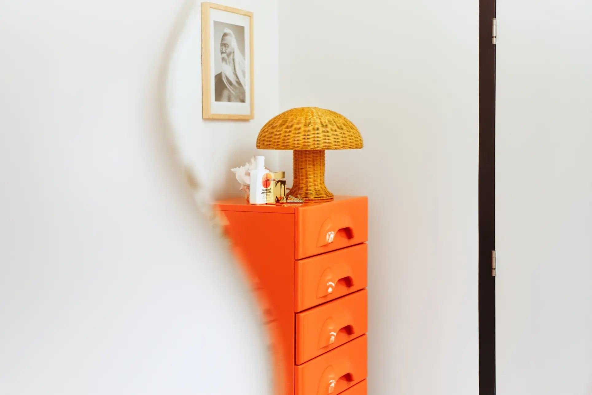 A modern room corner featuring an orange chest of drawers, the HKliving Rattan Table Lamp by HKliving, cozy mood lights, small decor items, and a framed black-and-white photo on a white wall. The image is partially blurred on the left side.