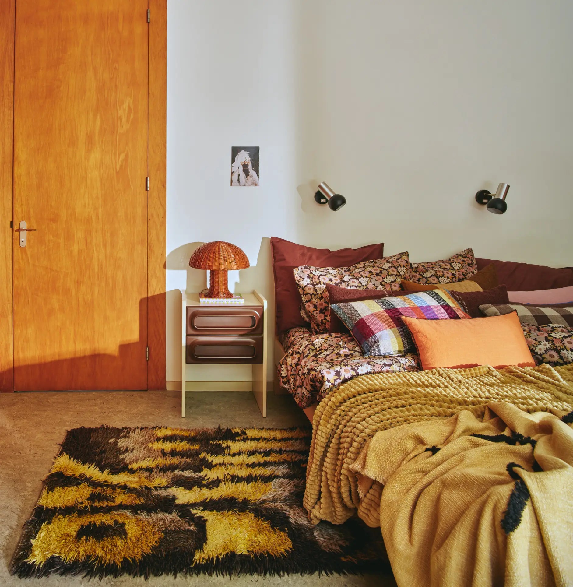 Cozy bedroom with patterned bedding, colorful pillows, and a mustard knit throw. A HKliving Rattan Table Lamp by HKliving sits on the nightstand. Country aesthetics are highlighted by a brown and yellow rug and cozy mood lights above the bed.