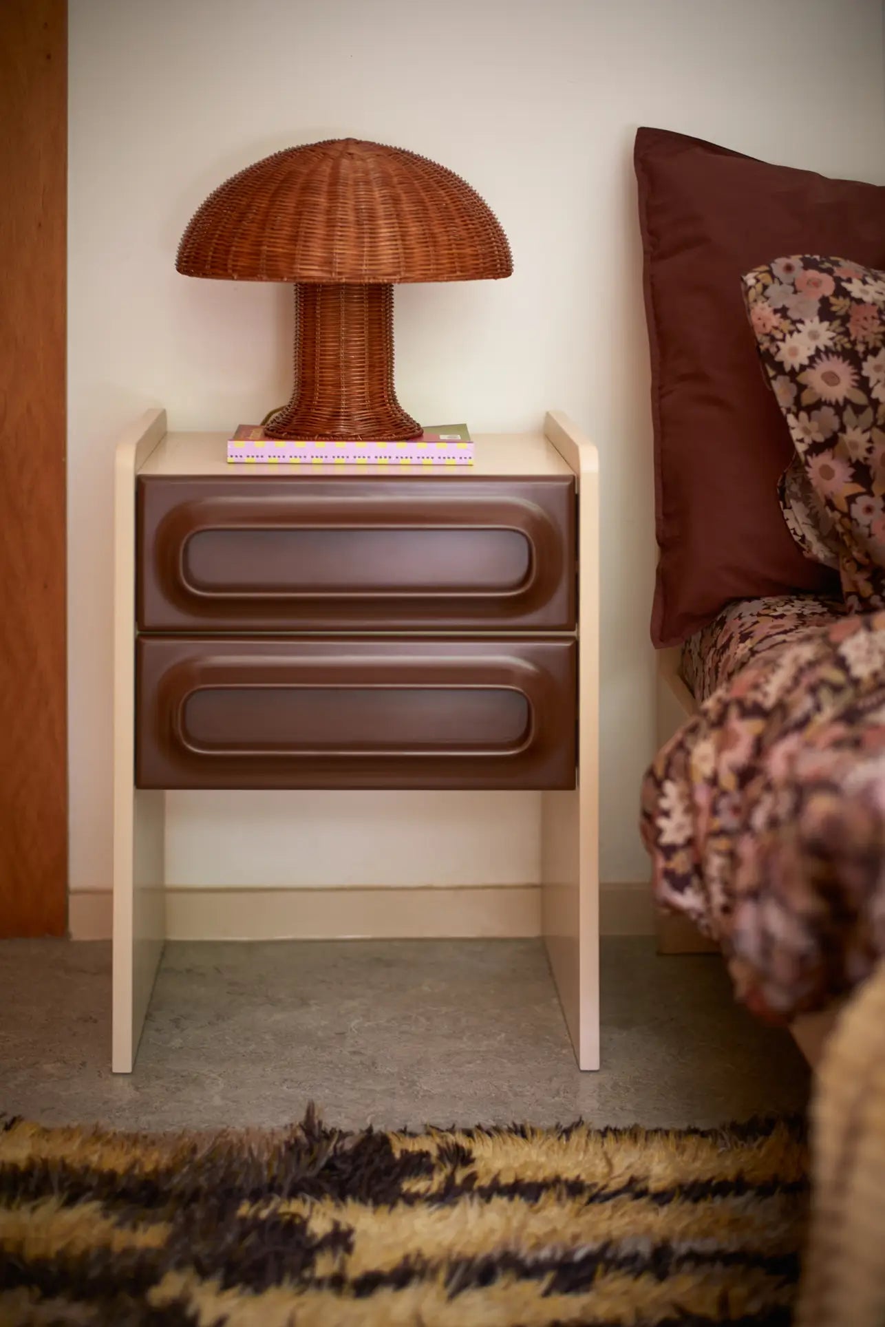 A modern nightstand with two brown drawers holds an HKliving Rattan Table Lamp, shaped like a mushroom, and a closed book. The adjacent bed features floral bedding, a burgundy pillow, and sits atop a patterned rug for a country look.