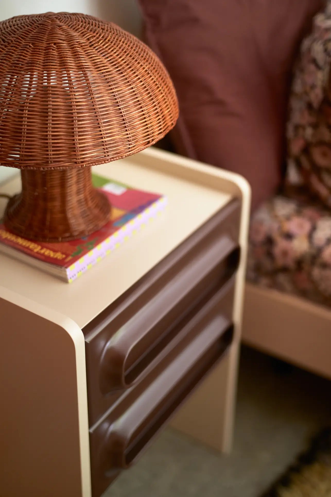 The HKliving Rattan Table Lamp, a charming rattan mushroom-shaped design from HKliving, rests on a modern nightstand with two brown drawers next to a pink-patterned bed and mauve pillow. A colorful book sits beneath this cozy country-style light.
