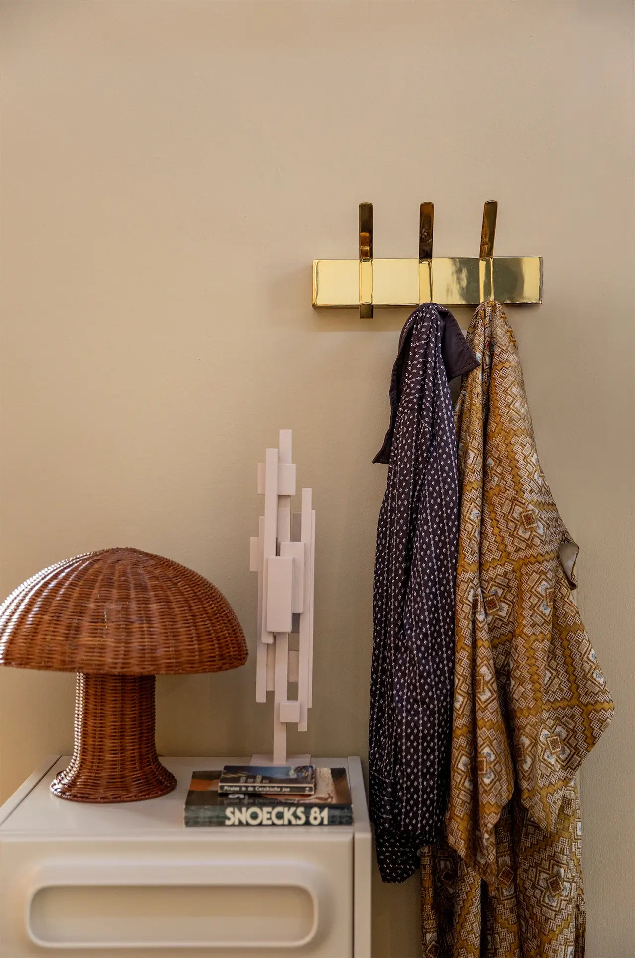 The HKliving Rattan Table Lamp, a white modern sculpture, and stacked books rest on a white table by a brass wall hook with two patterned scarves, creating cozy light and adding country style against the beige wall.