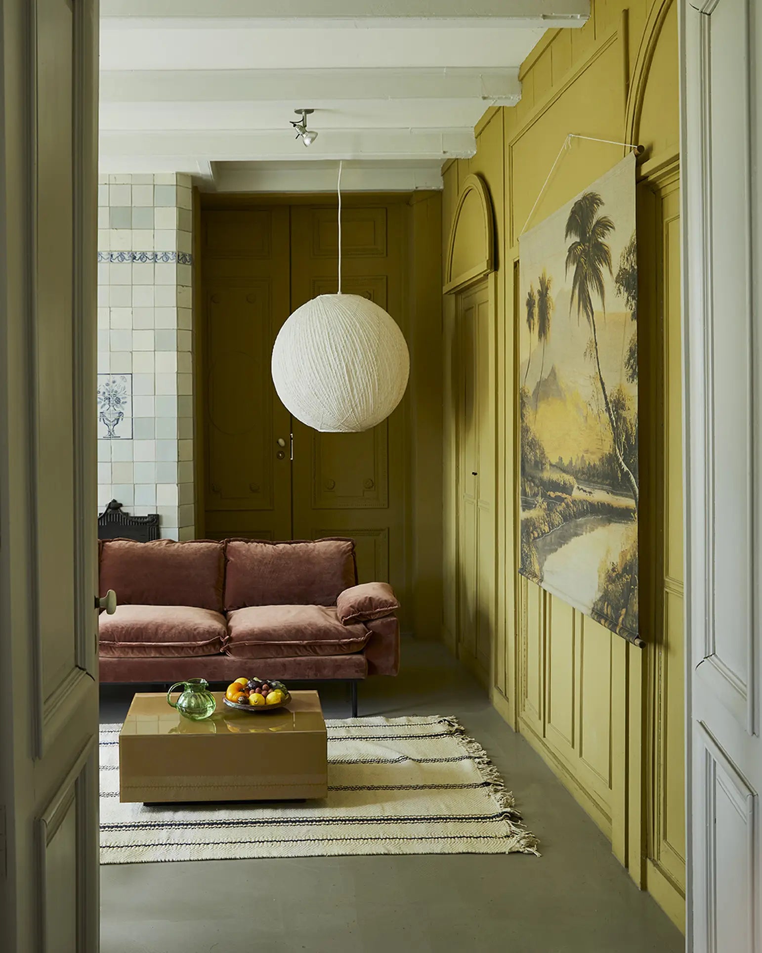 A cozy living space with mustard yellow paneled walls, the HKliving Retro Sofa, 4-Seats in pink velvet, a brown coffee table with fruit, a beige striped rug, a large round paper lantern, and a palm tree landscape painting.