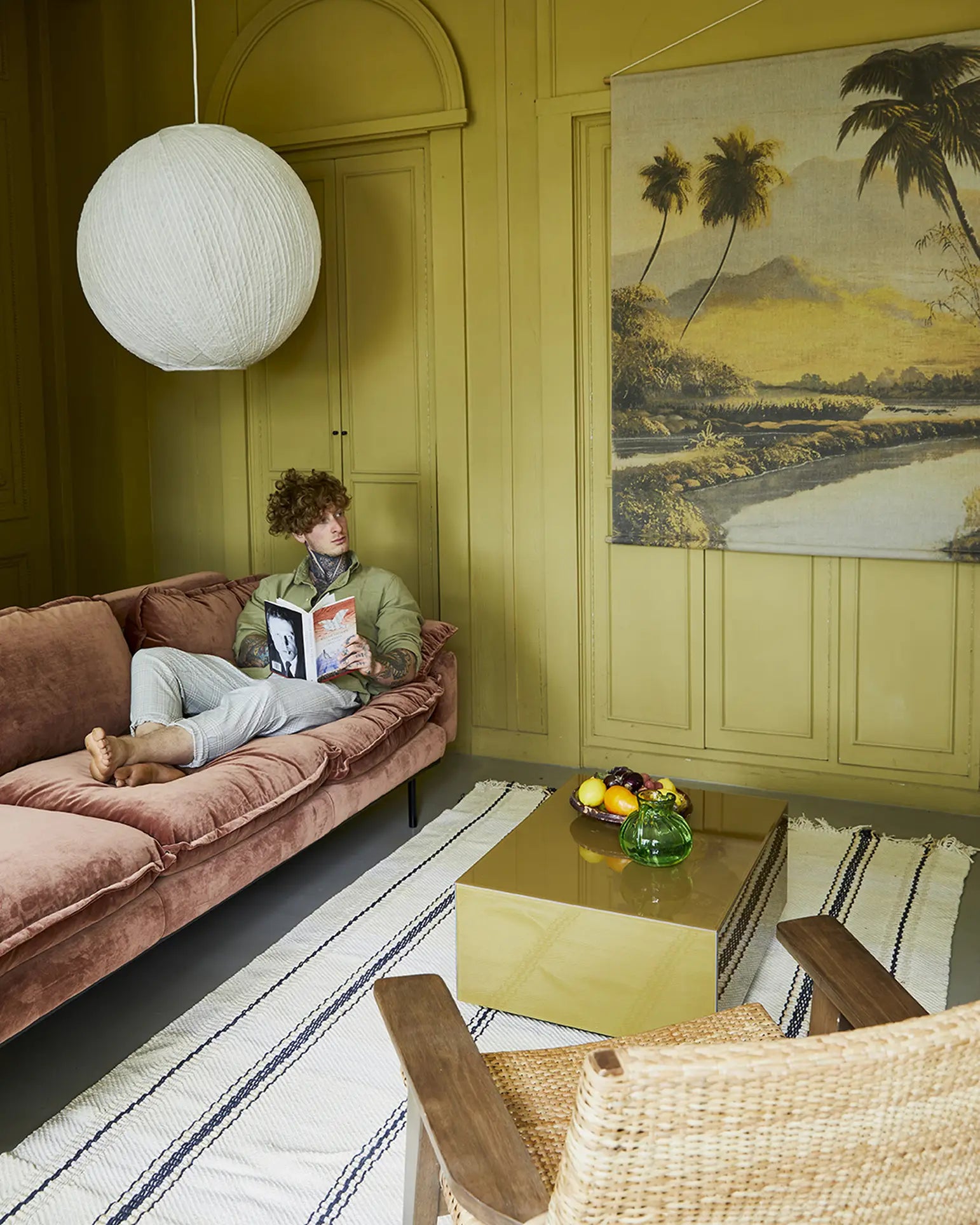 A person sits on the HKliving Retro Sofa, 4-Seats in a yellow-green living room with paneled walls, a round lamp, tropical wall art, a striped rug, and a gold coffee table topped with fruit and a vase.