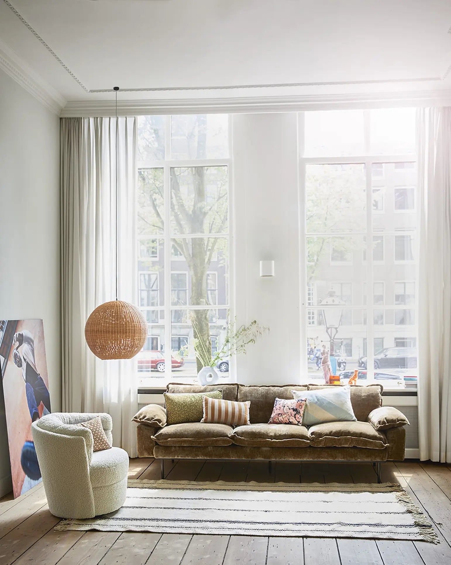 Bright living space with large windows, light curtains, the HKliving Retro Sofa 4-Seater in brown velvet with decorative pillows, a white plush armchair, striped rug, woven pendant light, and a large abstract artwork resting against the wall.
