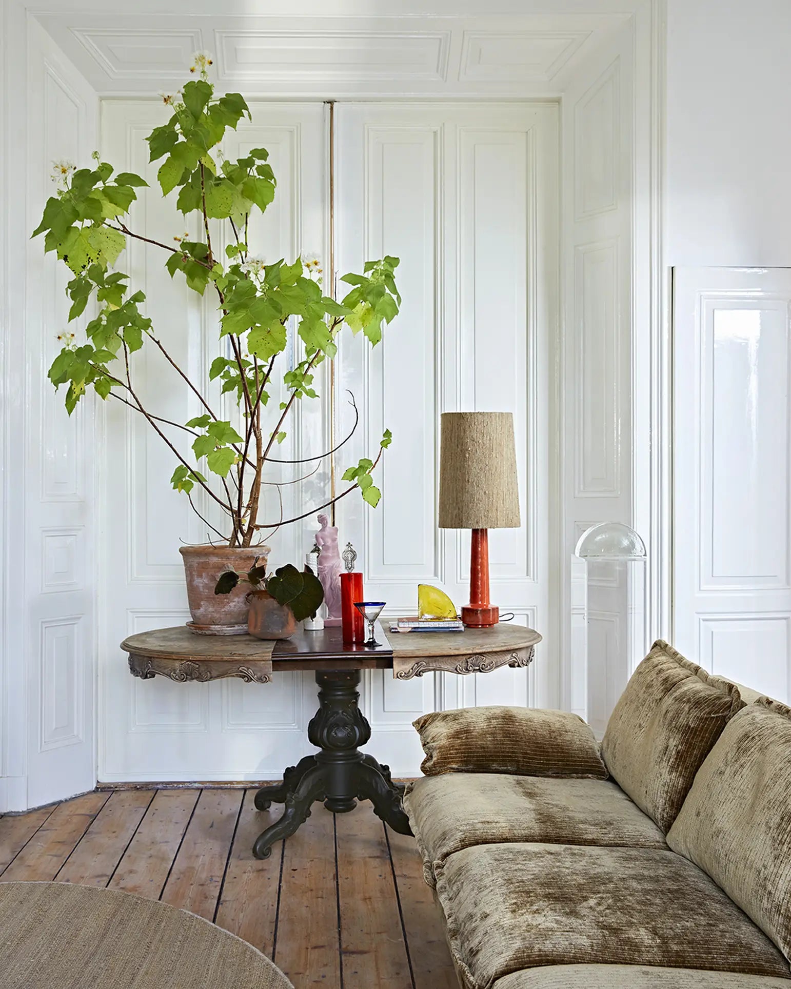 A cozy living space with an HKliving Retro Sofa, 4-Seats in velvet gold, wooden floors, a round table with a tall plant, lamp, red glassware, and decor, all framed by elegant white paneled walls.