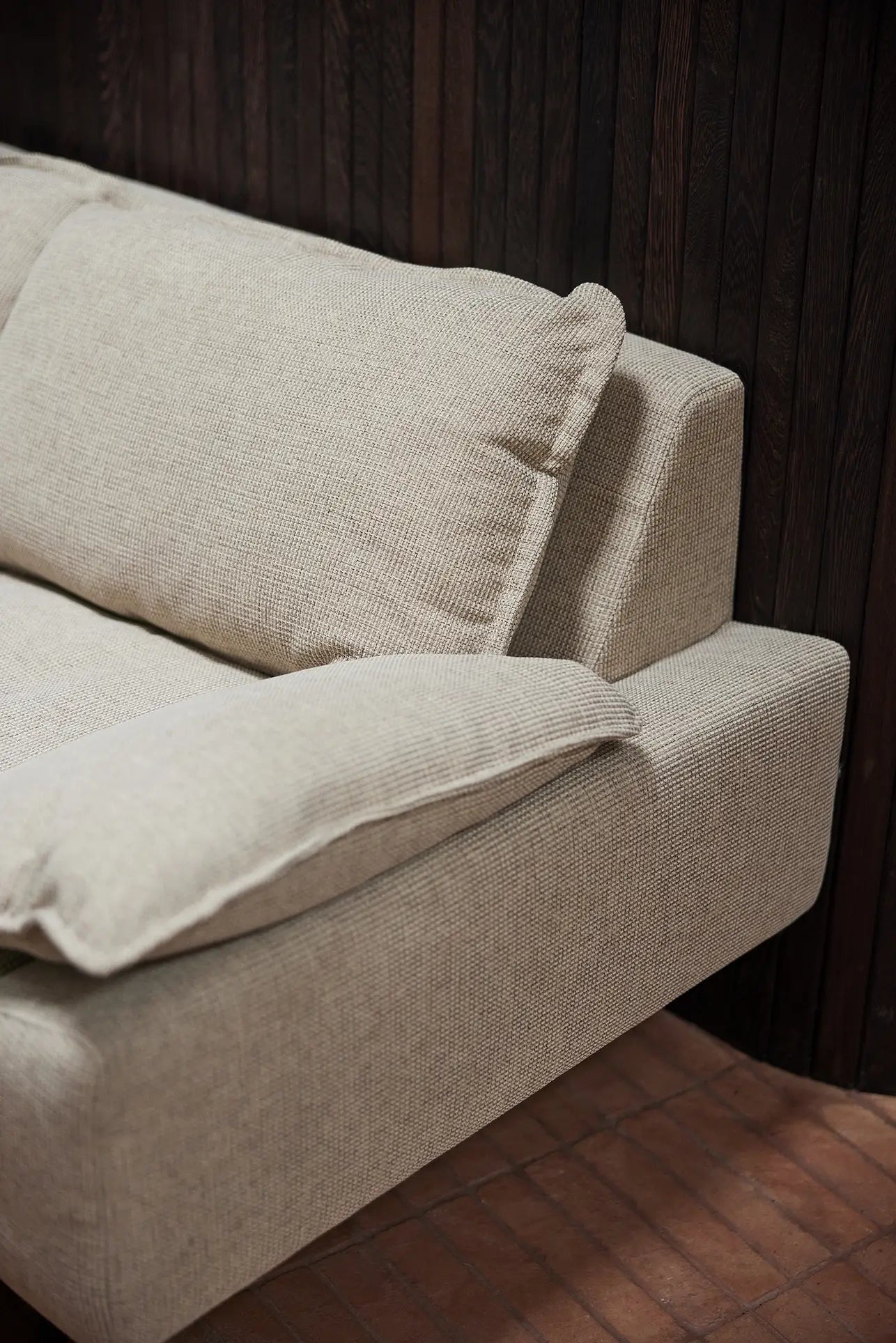 The HKliving Retro Sofa, 4-Seats by HKliving features large cushions and beige fabric for exceptional comfort, showcased against a dark wooden wall and reddish-brown tiled floor.