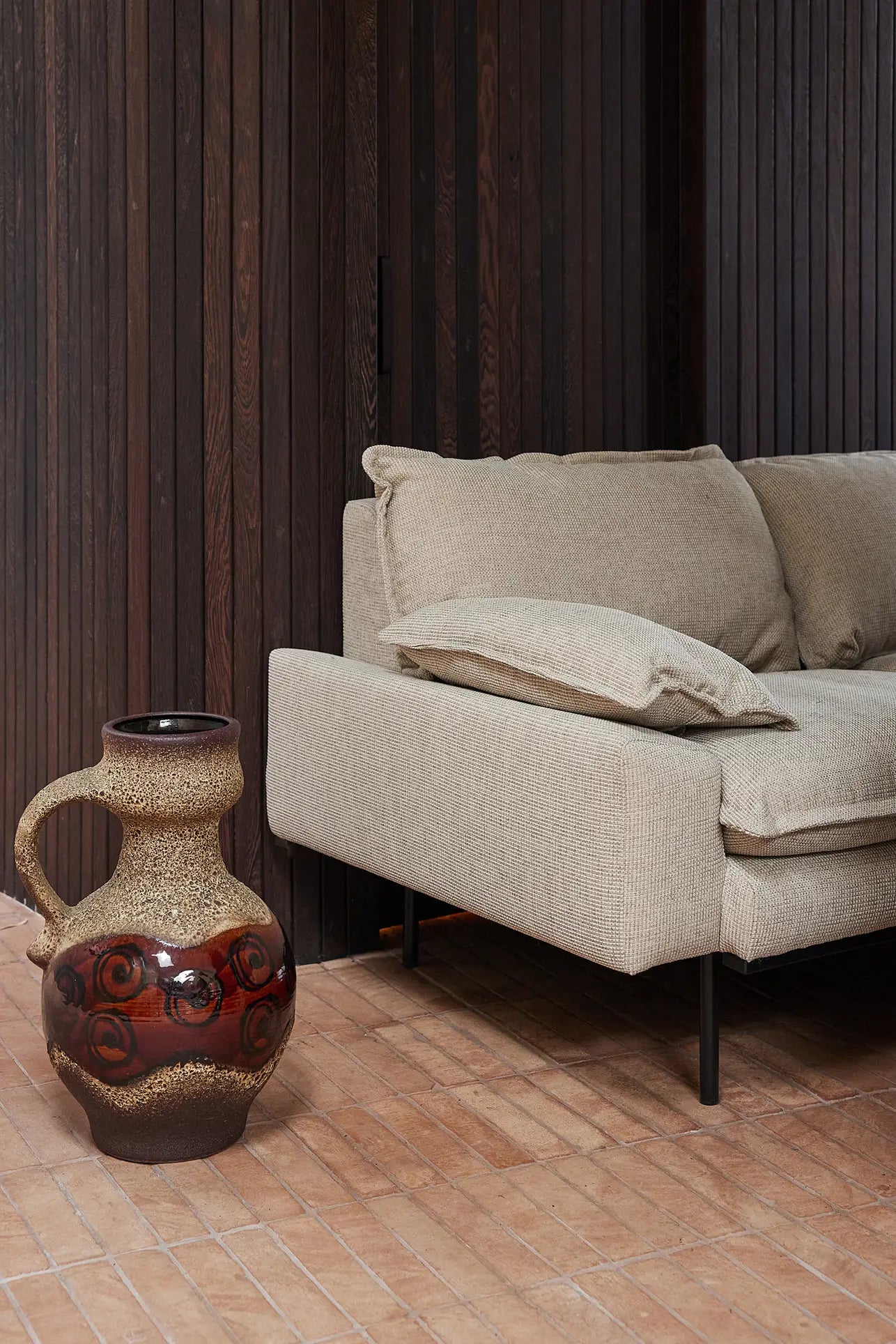 The HKliving Retro Sofa, 4-Seats in beige delivers comfort with plush cushions, set on a wooden floor next to a large ceramic vase adorned with brown and red patterns, all against rich dark wood paneling.