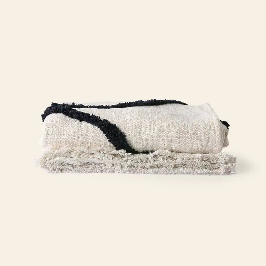 The HKliving Soft Woven Throw With Black Tufted Lines by HKliving is folded neatly on a light gray, fluffy rug, set against a plain beige background.